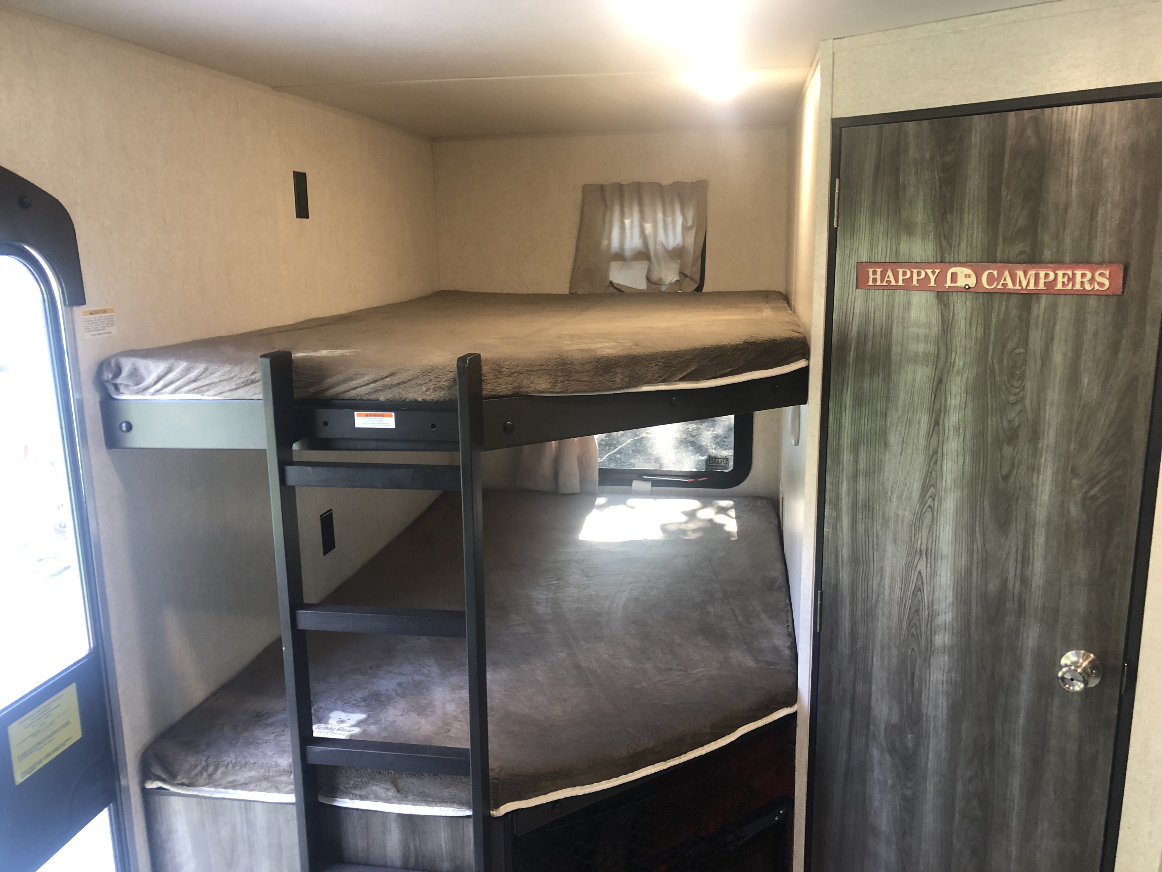 Inside an RV rental, showing bunk beds with cozy bedding. A window offers natural light, and a 'Happy Campers' sign adorns the door.. Jayco Highland Ridge 2019