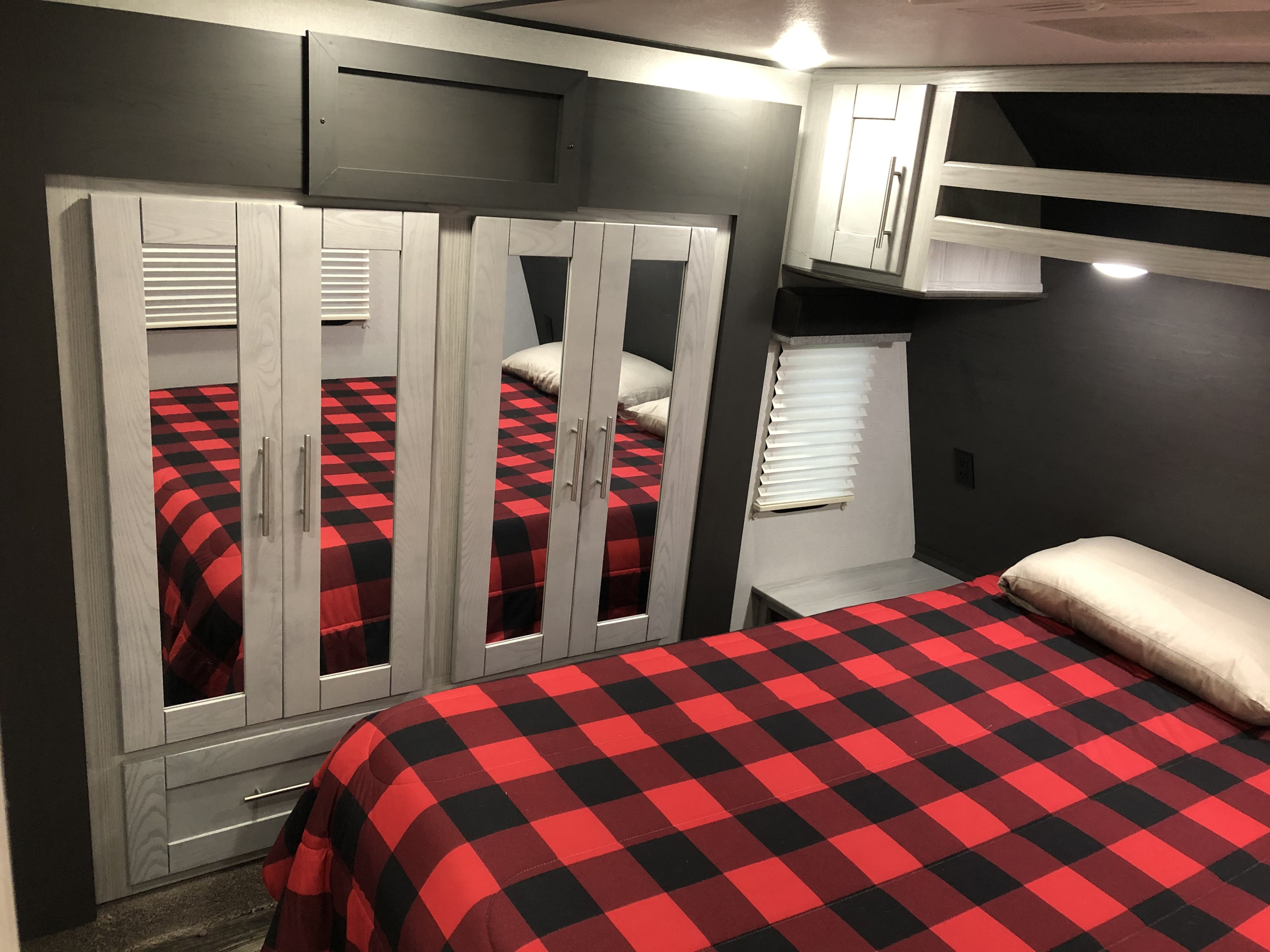 Inside an RV rental: a modern bedroom with a red & black plaid comforter, mirrored wardrobes, & overhead storage.. Keystone Fuzion 2021