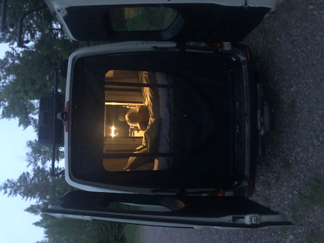 Cozy RV rental: Person relaxing in a brightly lit camper van bed at dusk with rear doors open.. Winnebago Revel 2020