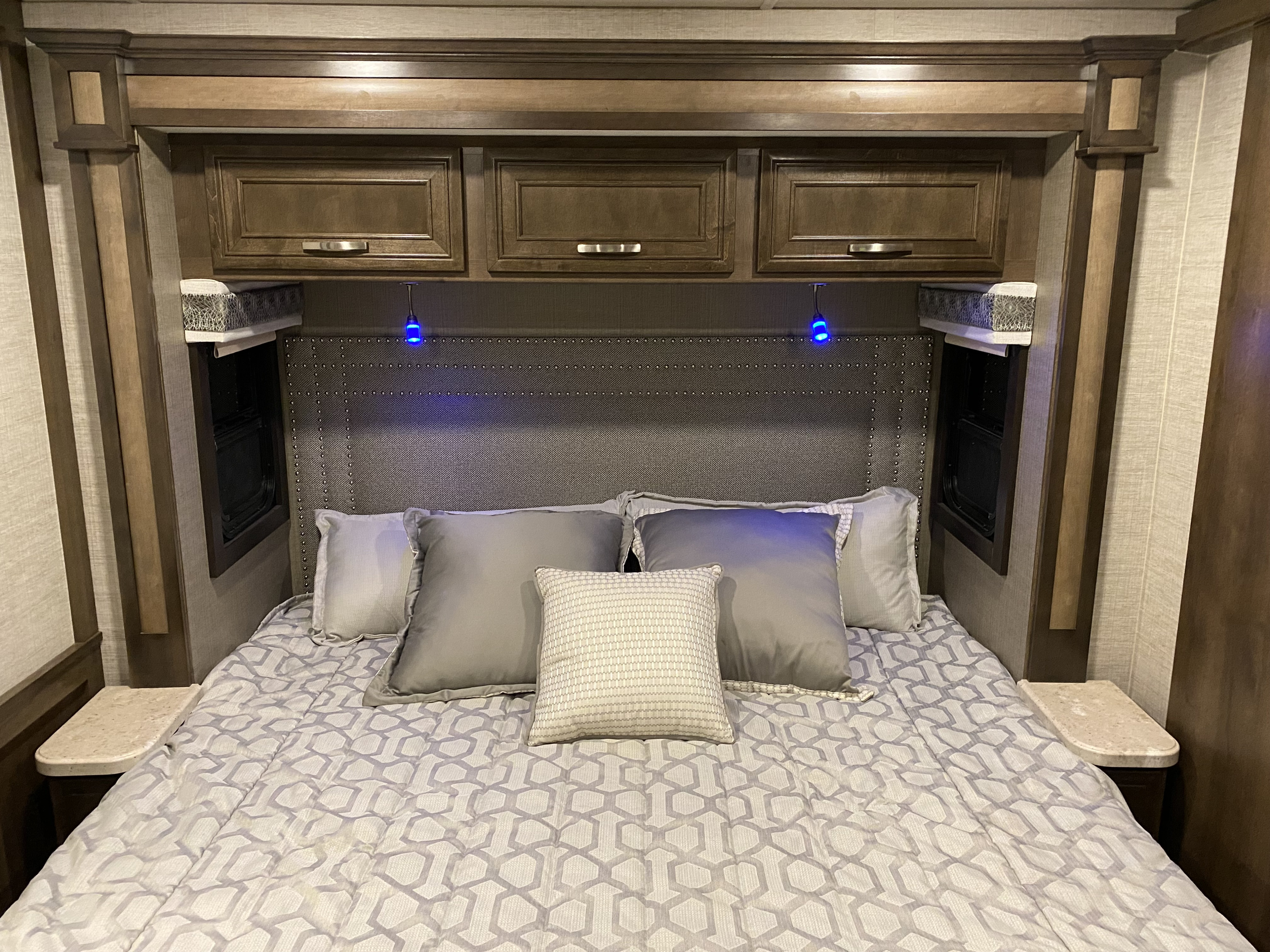 This cozy RV rental bedroom features a bed with geometric comforter, gray pillows, wooden cabinets, and blue headboard lights.. Entegra Coach Vision XL 2021