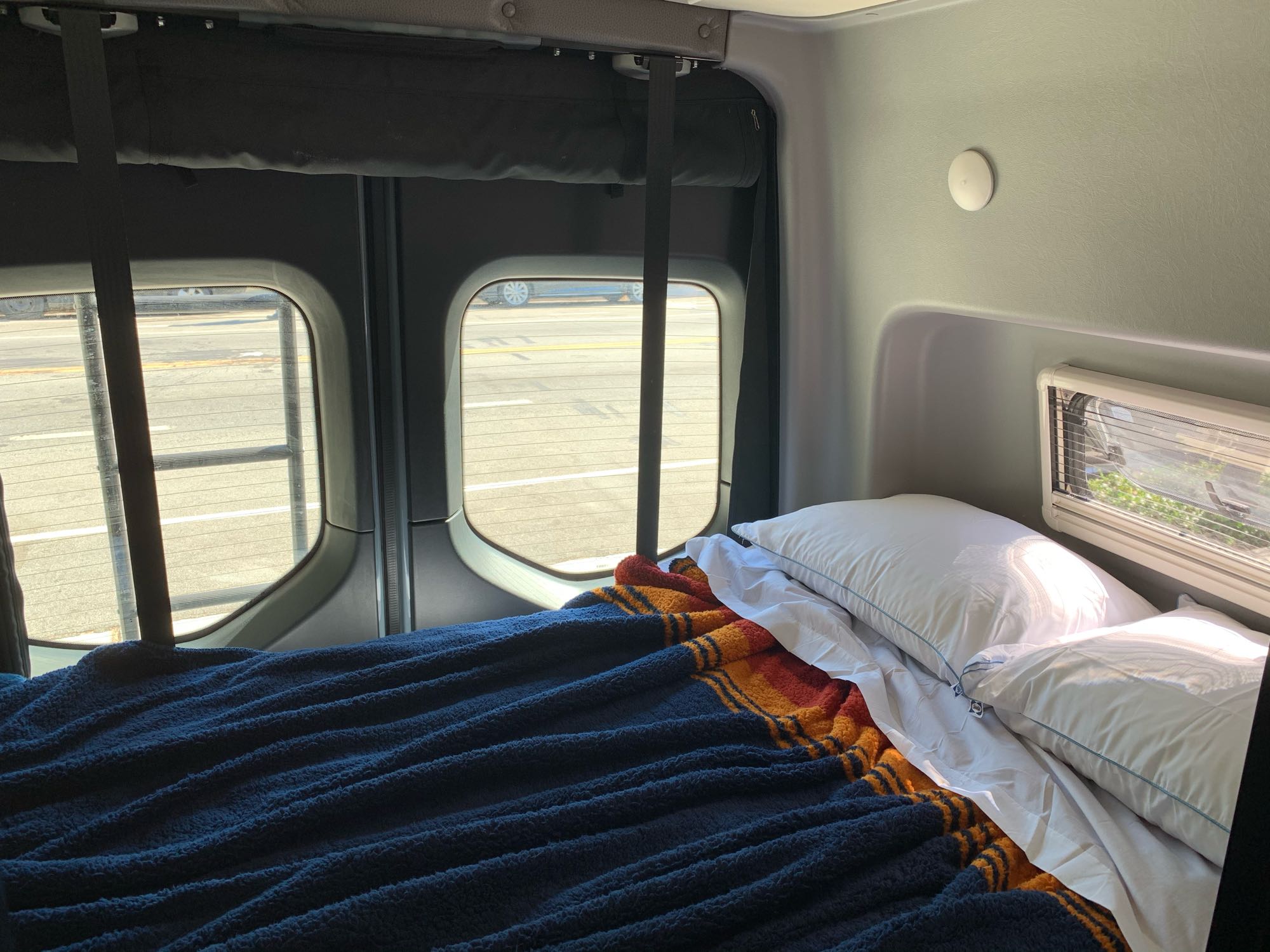 Interior of an RV rental, featuring a made bed with pillows, blankets, and large windows revealing a sunlit street outside.. Winnebago Revel 2020