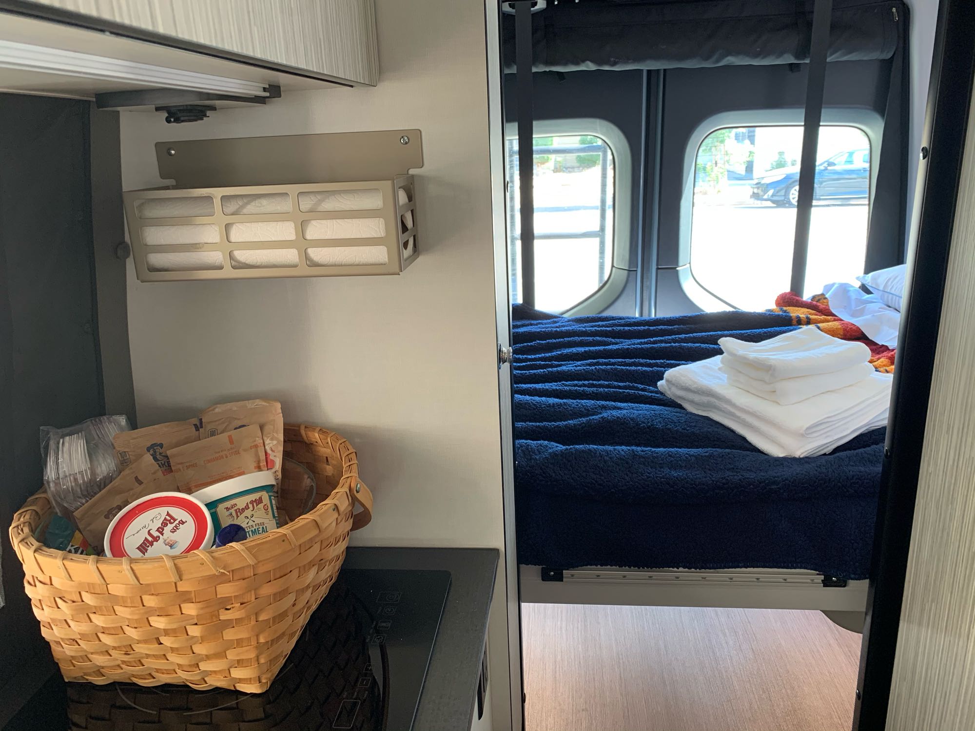 Inside this RV rental: kitchen counter with food basket, paper towels, and a cozy bed with blankets & towels.. Winnebago Revel 2020