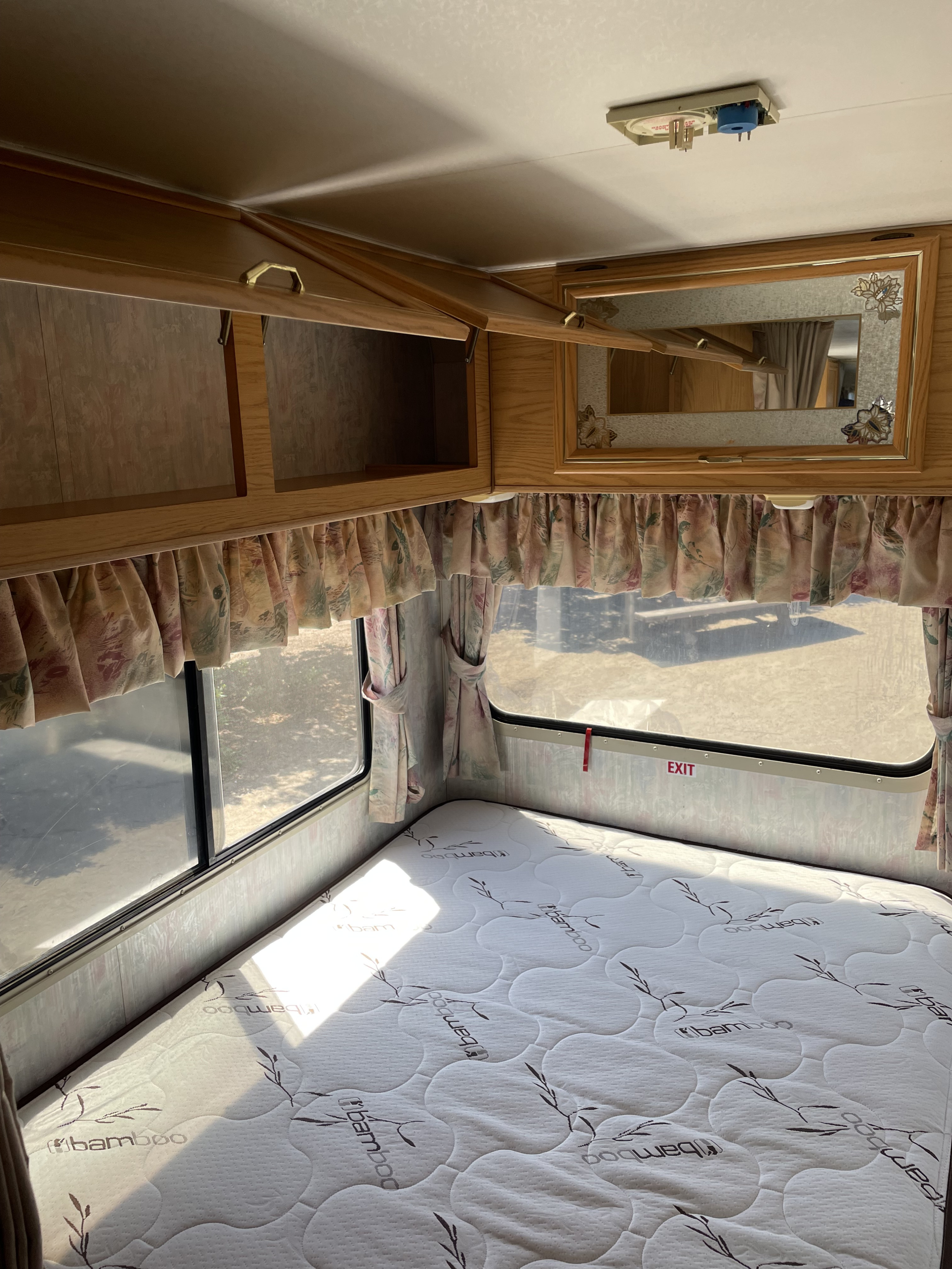 Bright RV rental bedroom: a "Bamboo" mattress, open wood cabinets, windows with floral curtains, and a mirror.. Itasca Spirit 1995