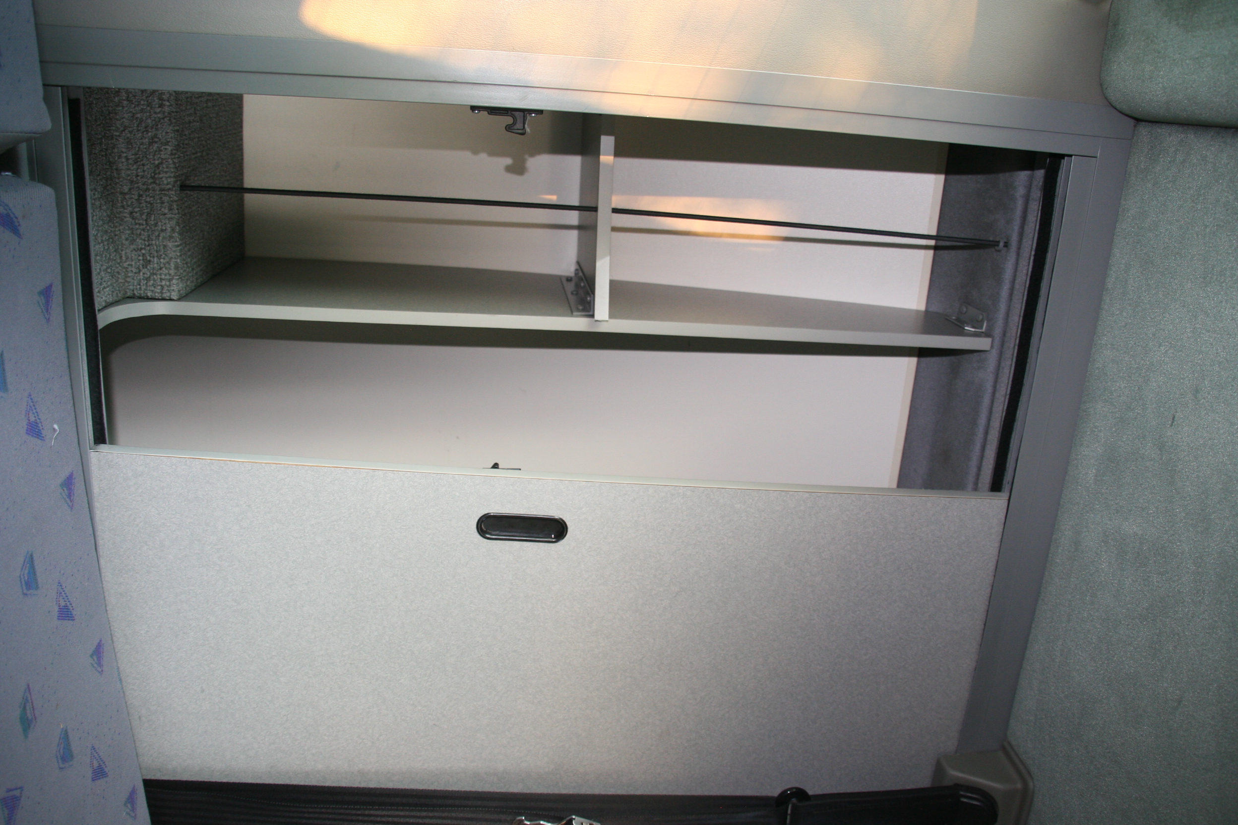 Open overhead storage with shelves and a latch in an RV rental interior, showing textured walls and patterned seating.. Volkswagen T4 2002