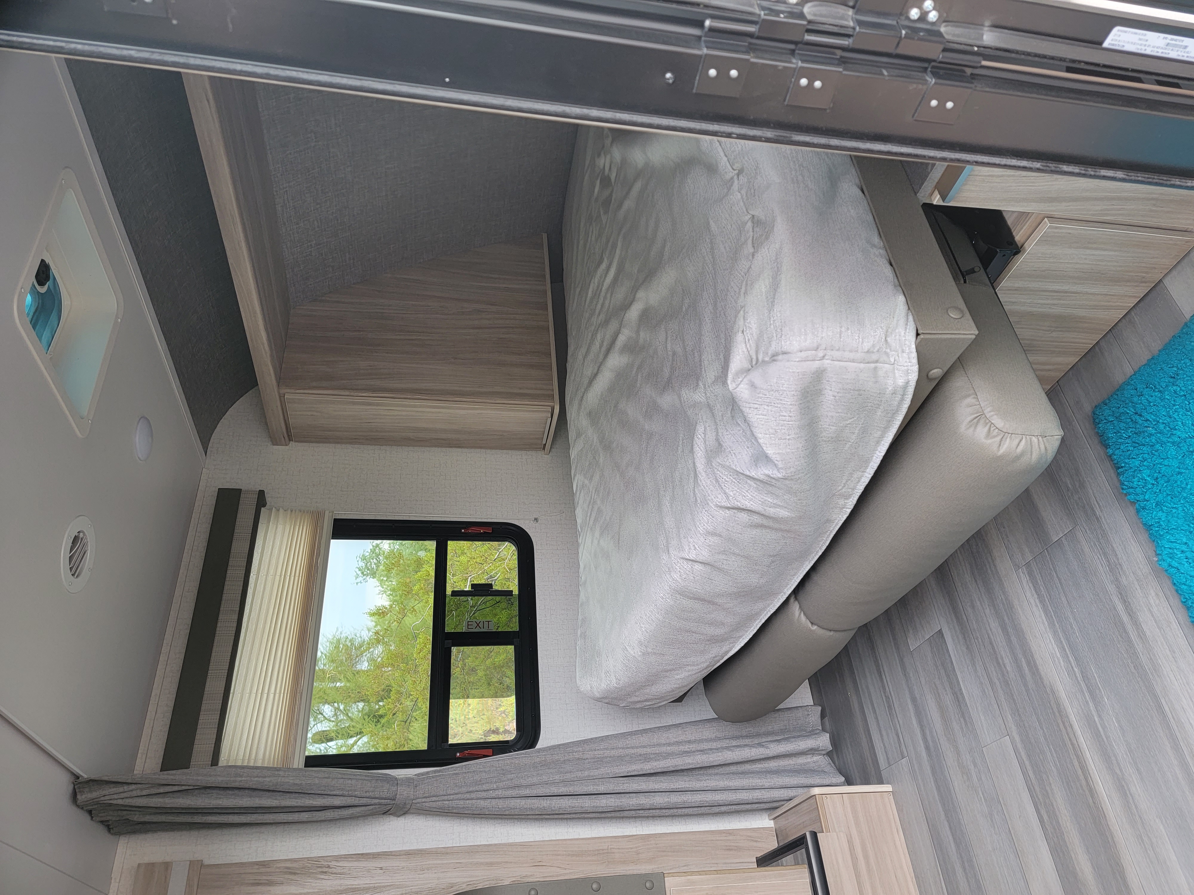 RV rental bedroom with a light gray bed, wooden overhead cabinets, and a window displaying an 'EXIT' sign. Features sleek, gray flooring.. Winnebago Micro Minnie 2021