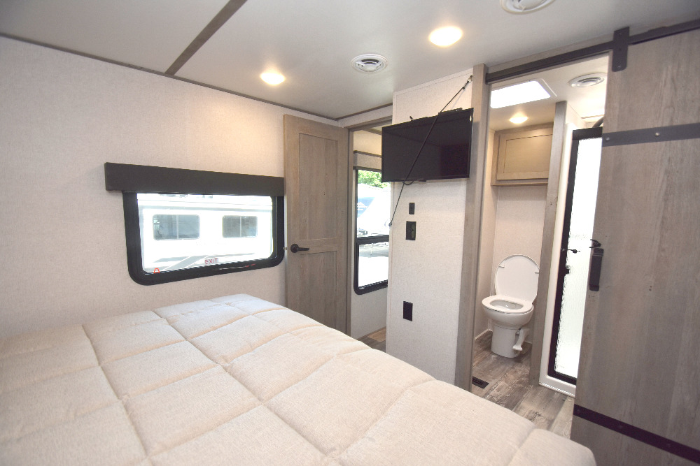 Modern RV rental interior: bedroom with bed, TV, and nearby bathroom with toilet & barn door.. Keystone Raptor 2023