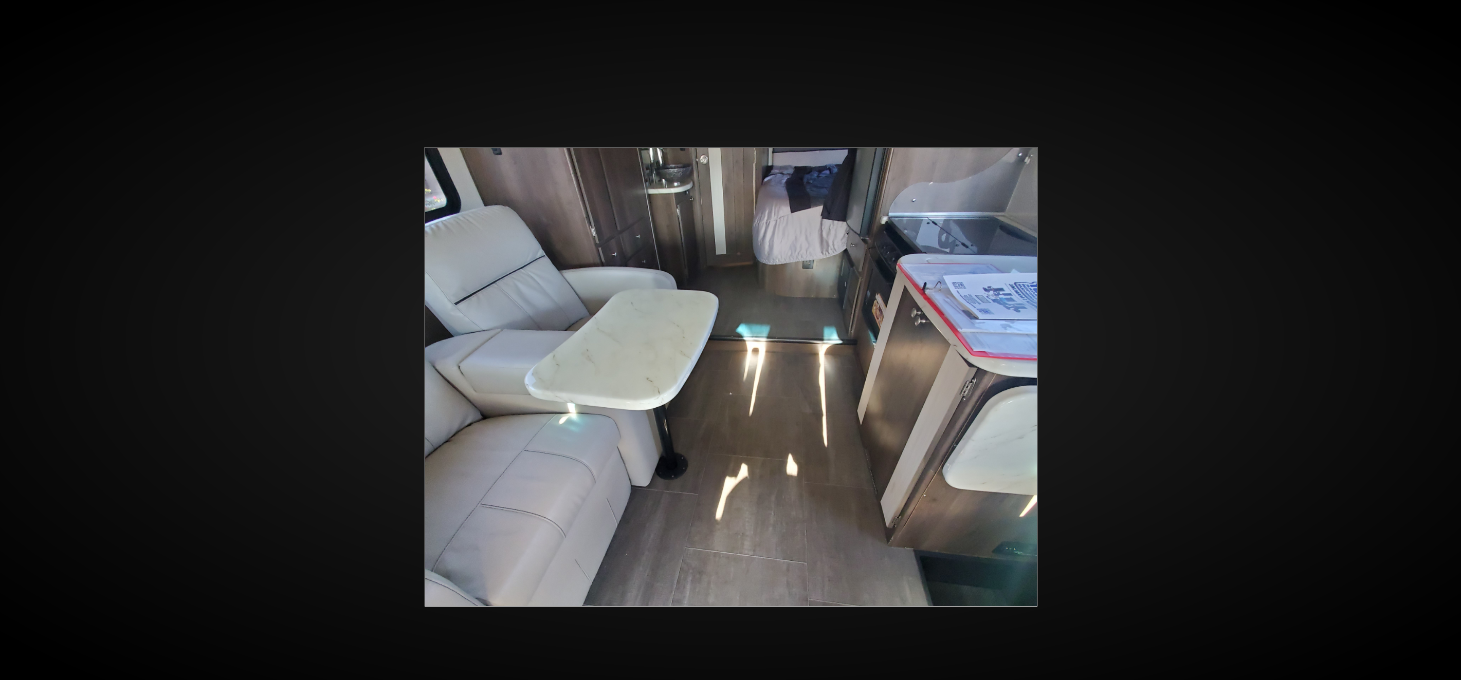RV rental interior featuring cream recliners, a small table, kitchen counter, and bedroom beyond. Sunlight streams on the floor.. Coachmen Prism 2018