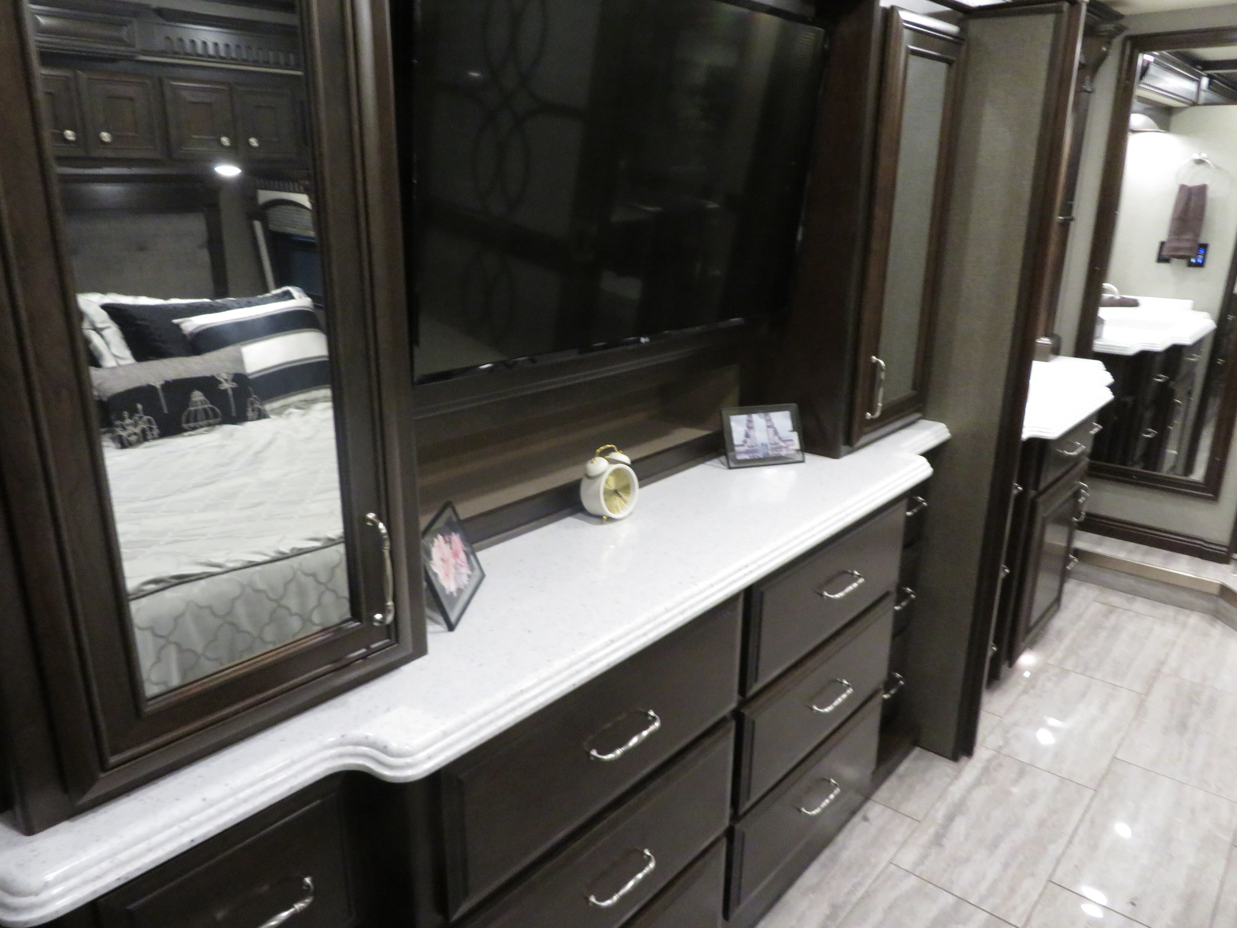 Spacious RV rental interior featuring a bedroom area with a dresser, TV, and reflected bed, plus a modern vanity leading to a bathroom.. Monaco Signature 2019