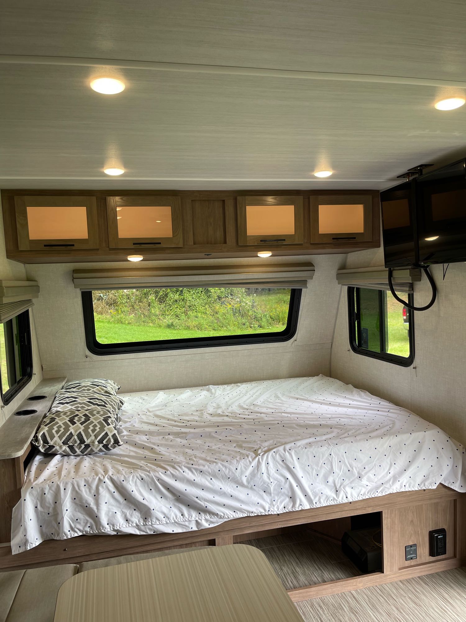 Interior of an RV rental bedroom showing a bed, overhead cabinets, and windows with green outdoor views.. No Boundaries 19 2021