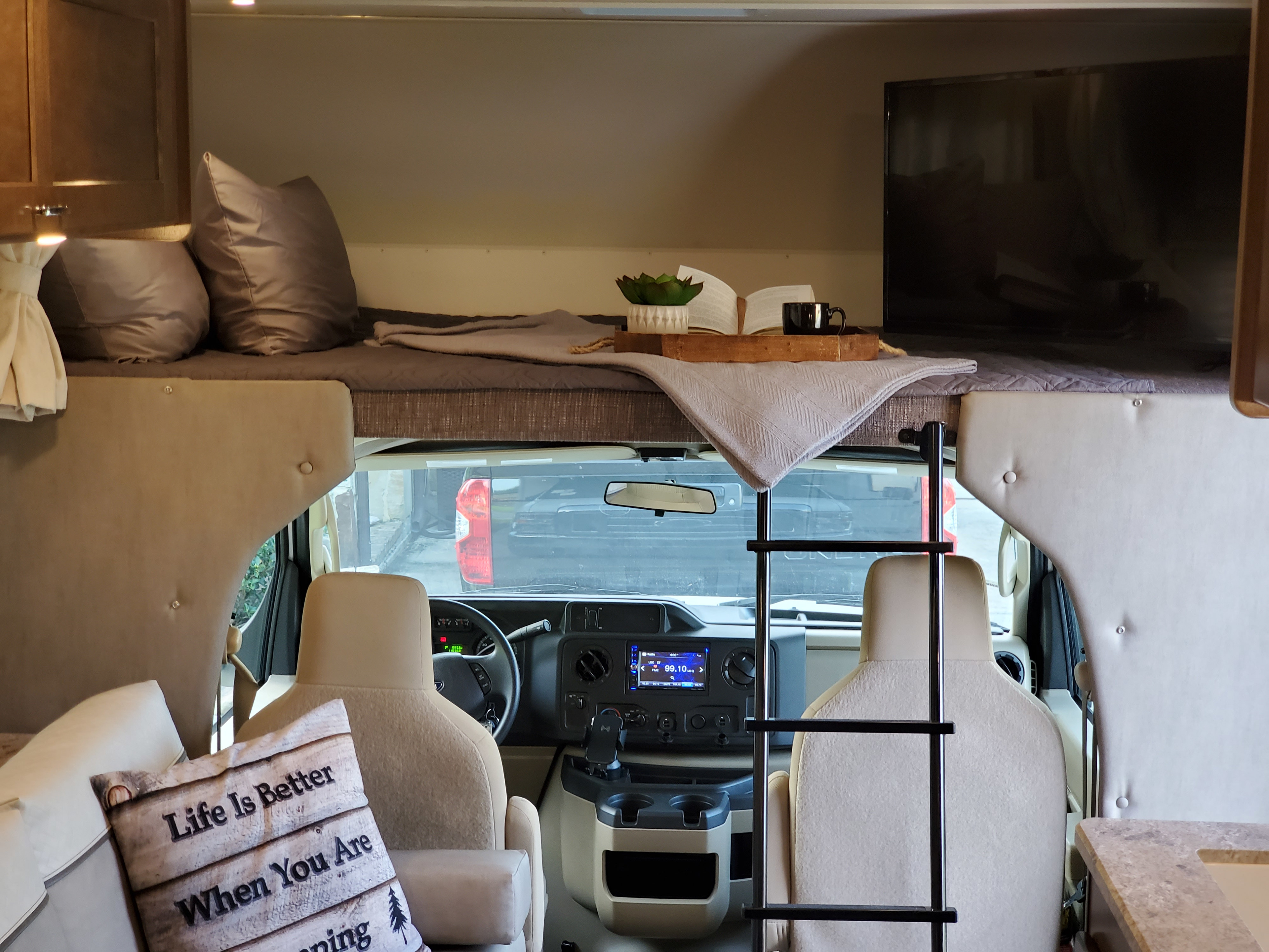 RV rental interior: overhead bunk with TV & reading nook, ladder, front cab, and a "Camping" pillow.. Winnebago Outlook 2019