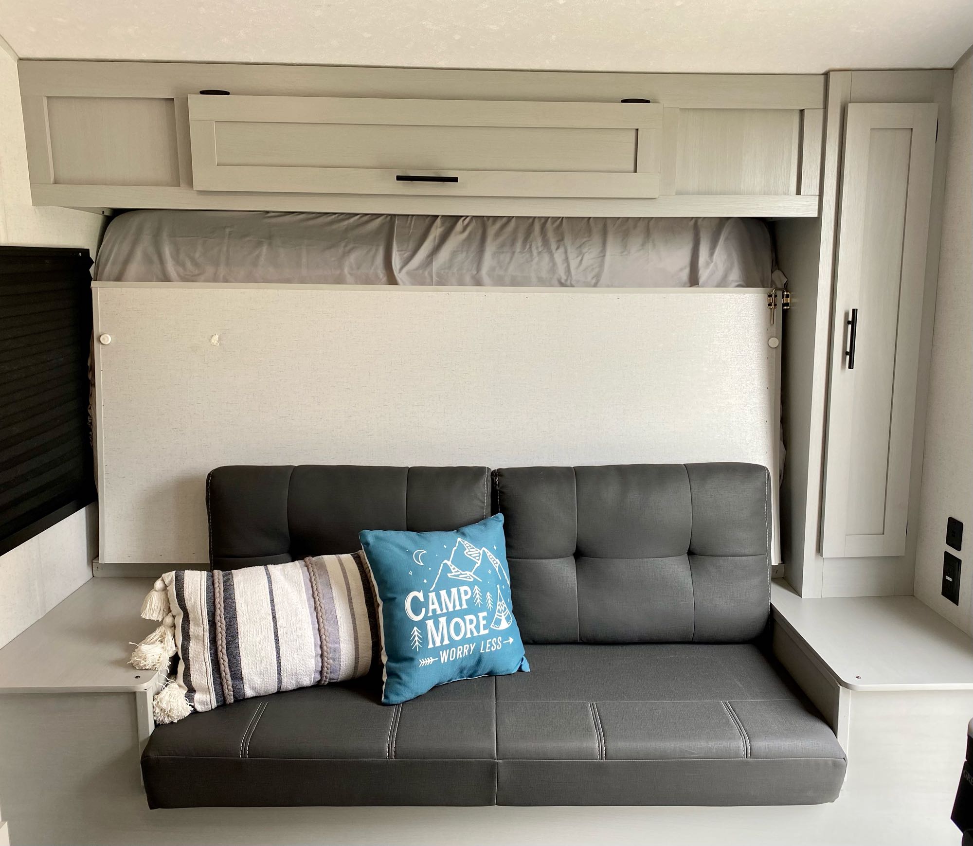 Inside an RV rental: a gray sofa below a stowed murphy bed, surrounded by light cabinets and shelving.. Forest River Wildwood 2021