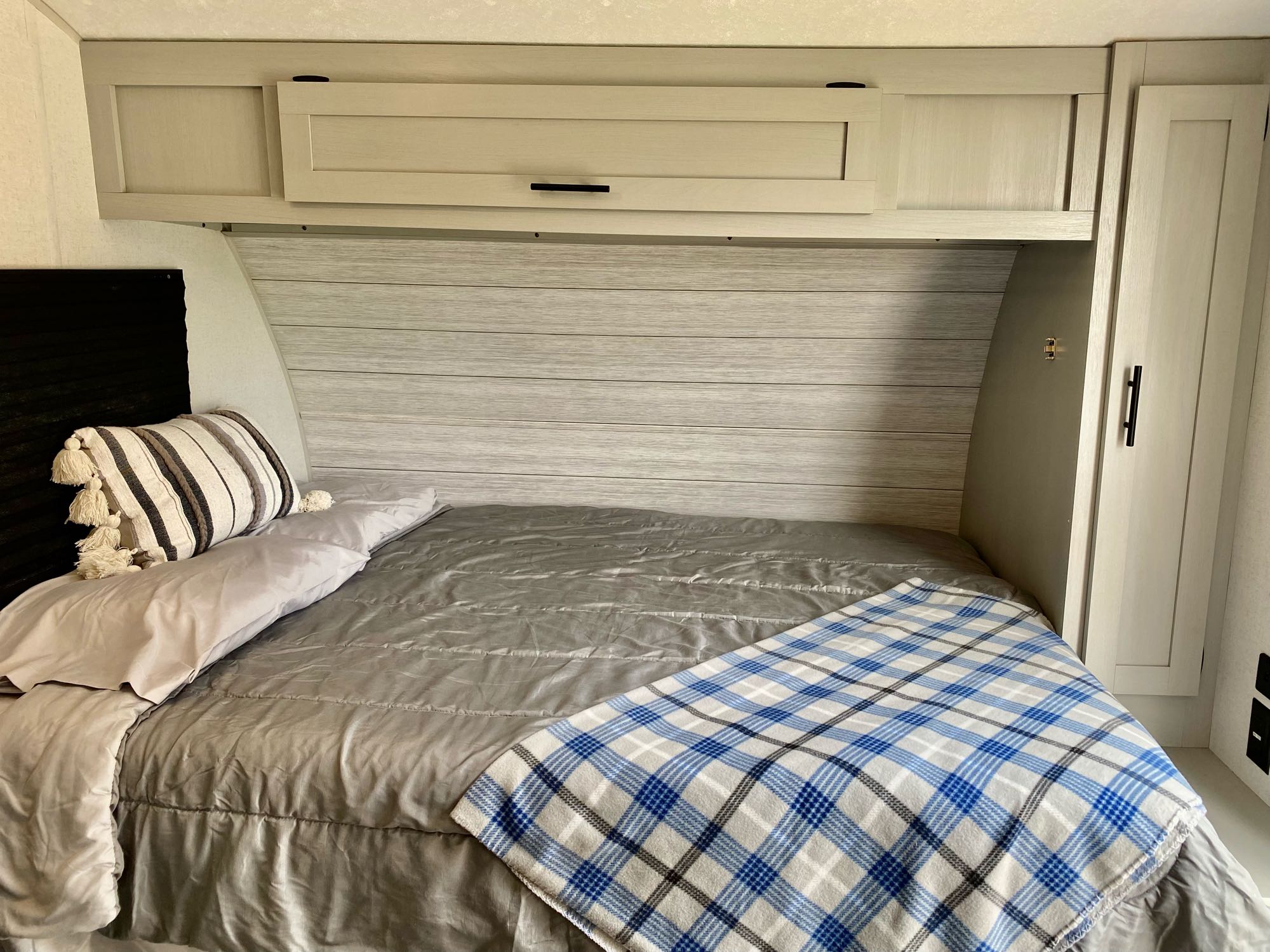 Cozy RV rental bedroom with light wood interior, overhead cabinets, and a neatly made bed with plaid blanket.. Forest River Wildwood 2021