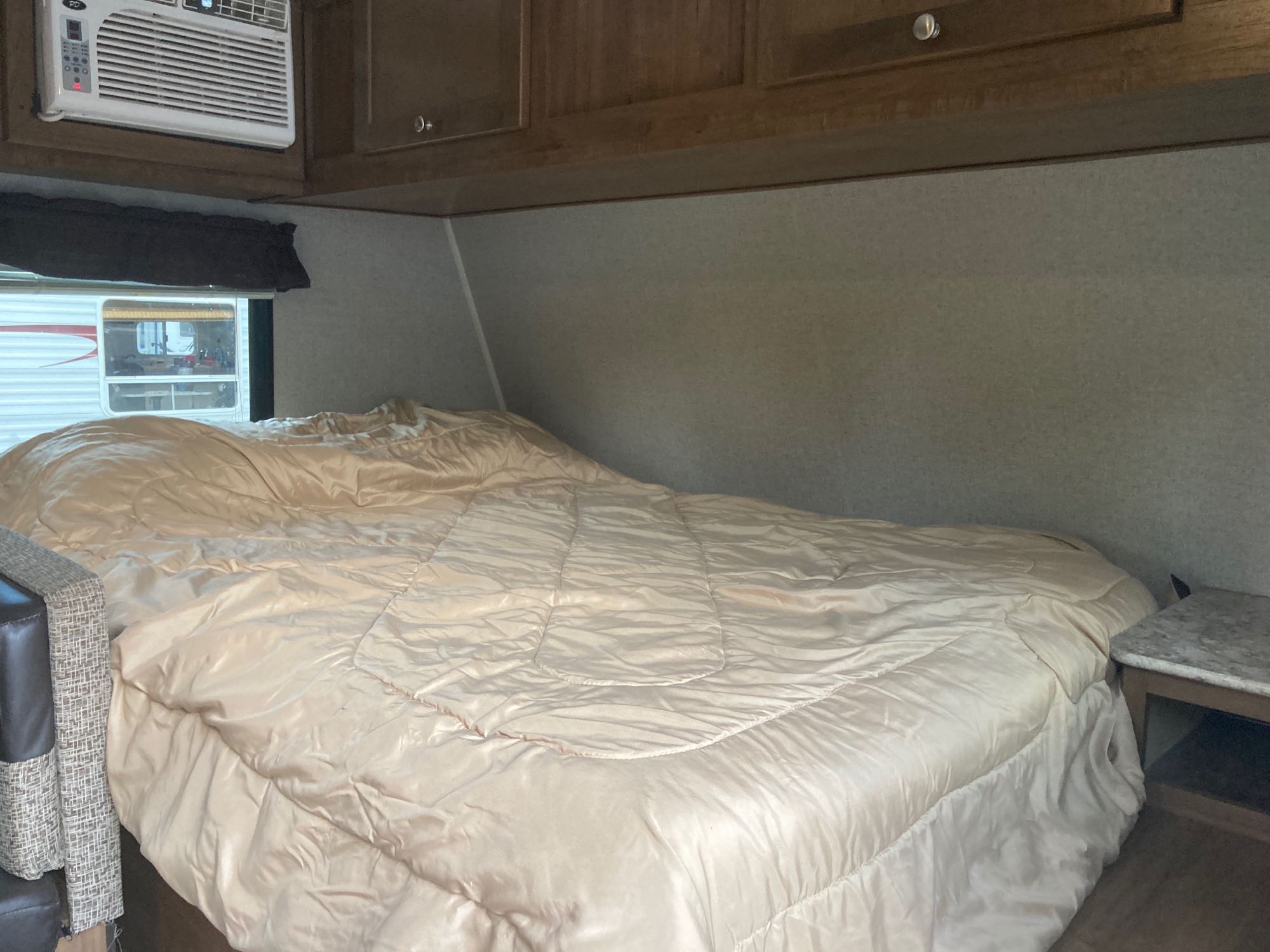 **RV rental bedroom interior**: Features a large bed with a light comforter, overhead cabinets, a mounted AC unit, and a window showing another RV.. Dutchmen Aspen Trail 2018