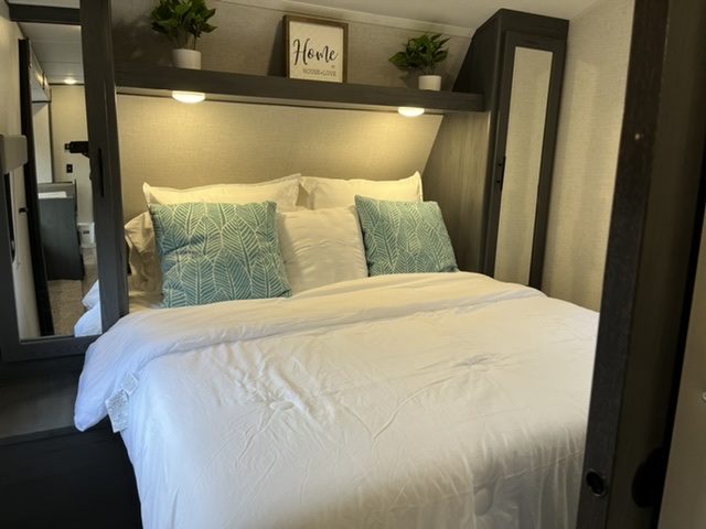 Cozy RV rental bedroom featuring a neatly made queen bed, decorative pillows, overhead lighting, and storage. Clean and inviting.. Dutchmen Aspen Trail 2021