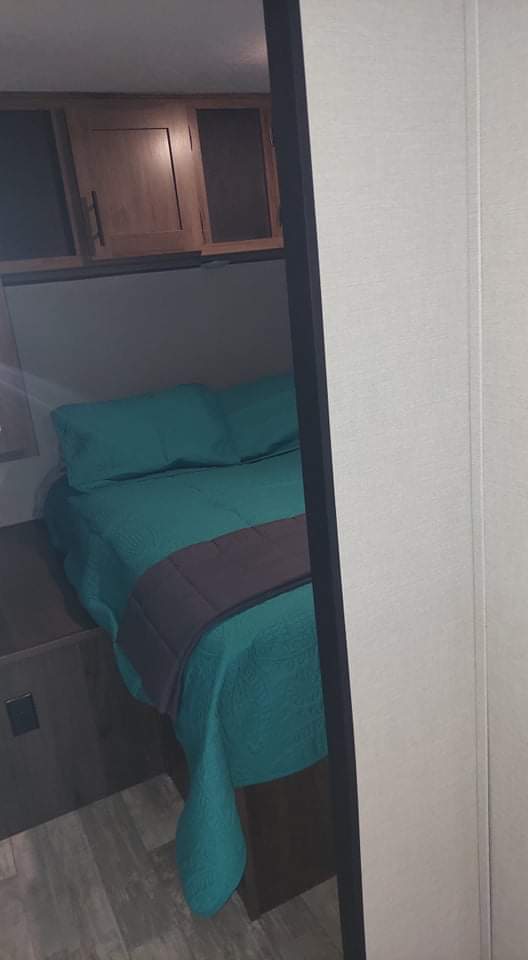 Cozy RV rental bedroom features a teal bed, overhead cabinets, and wood-look flooring.. Prime Time Avenger 2021