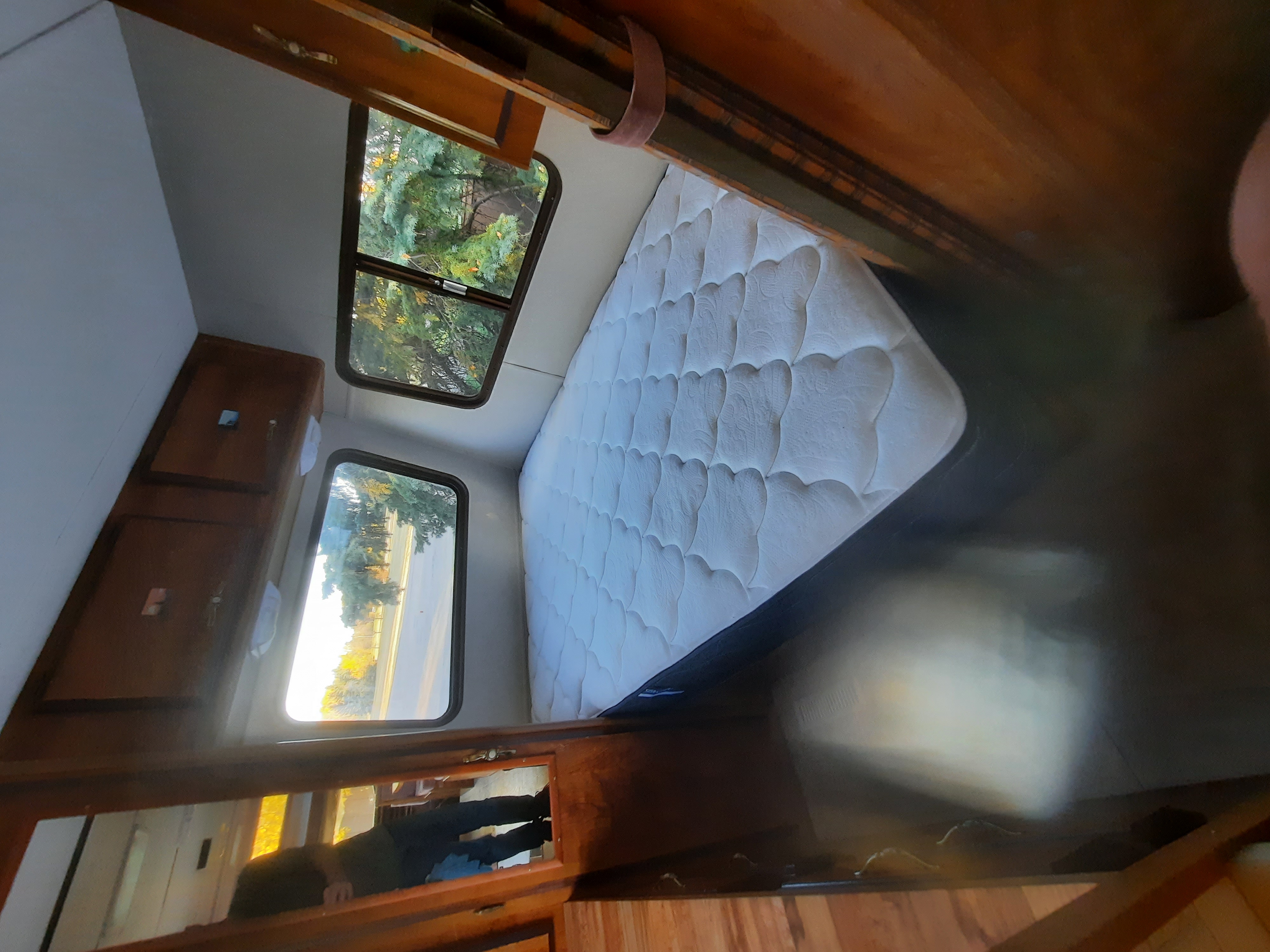 Inside an RV rental, a white quilted mattress sits by two windows overlooking trees and road. Wooden cabinets line the wall.. Ford Econoline 1986