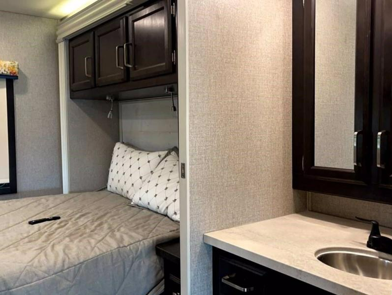 Inside an RV rental: a cozy bedroom with overhead storage, reading lights, and a nearby bathroom vanity with sink.. Jayco Greyhawk 2021