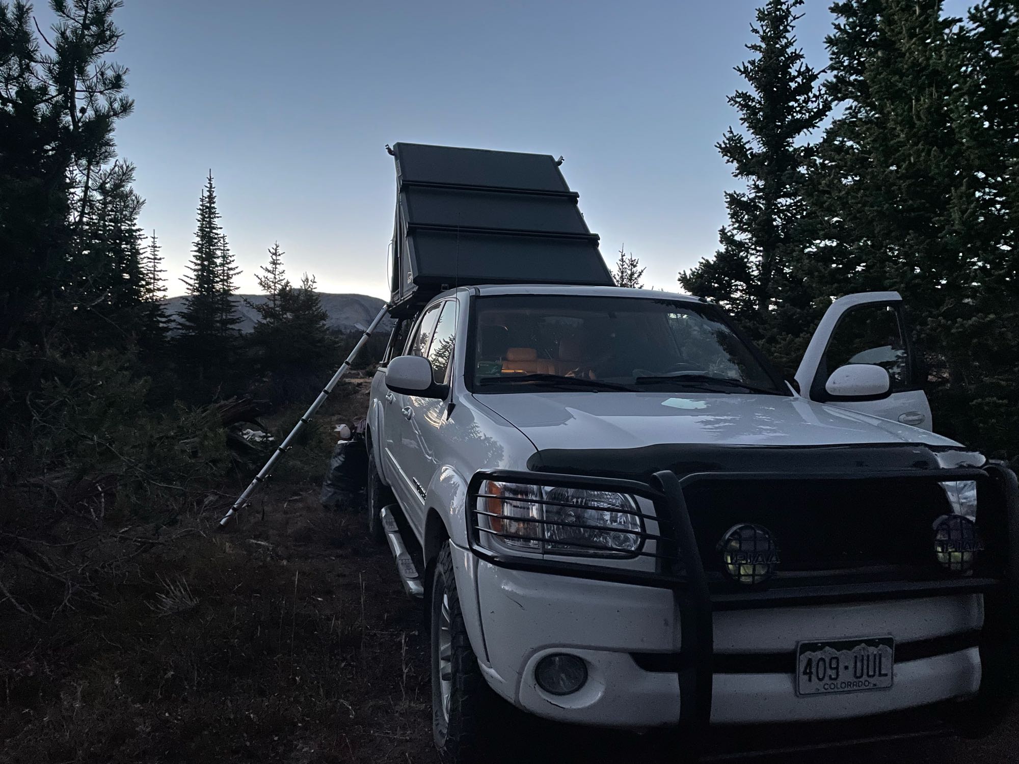 White overland truck with a rooftop tent, perfect for an RV rental adventure, parked amidst evergreen trees at twilight.. American Dream Toyota 2006