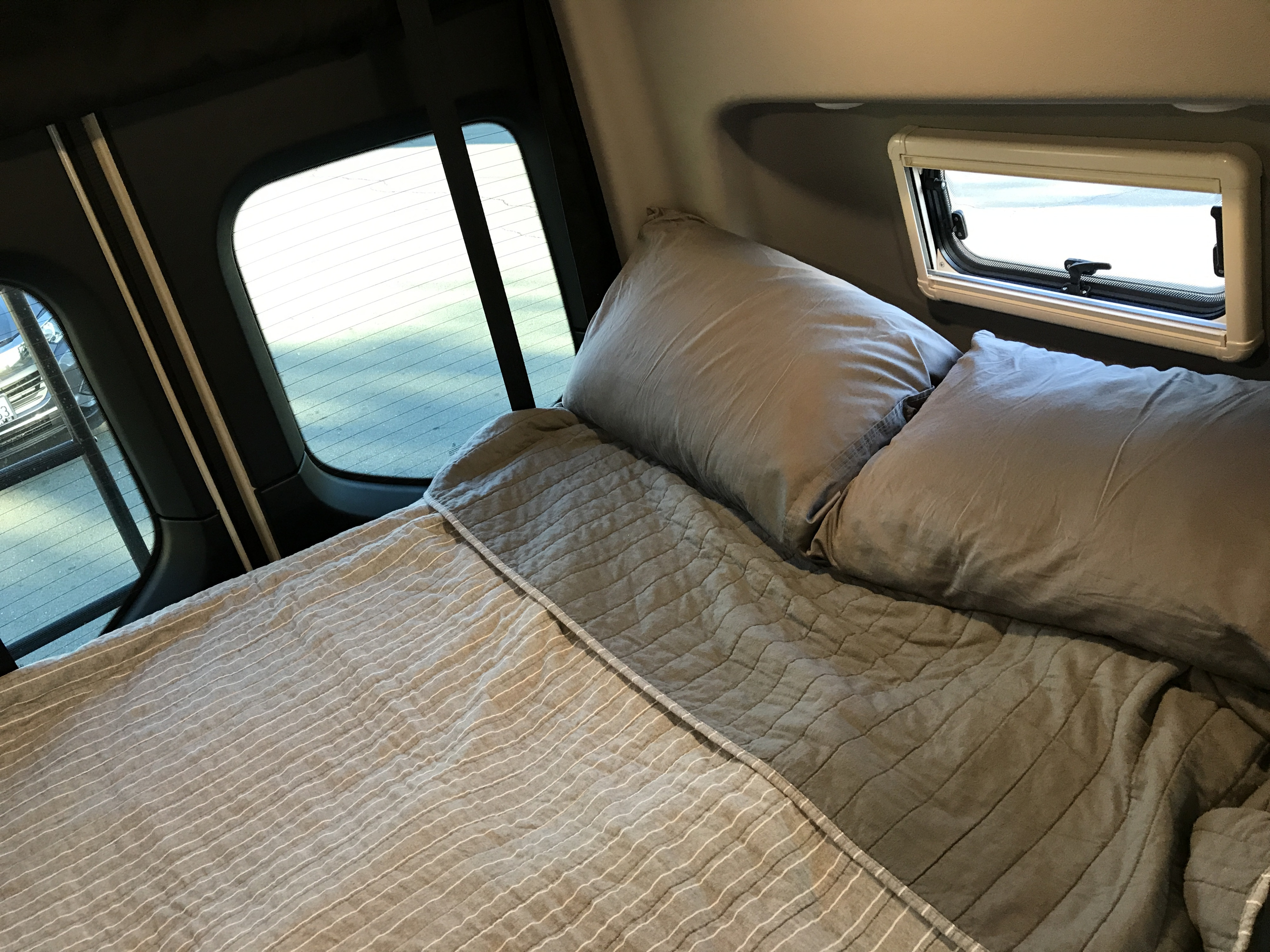 This RV rental shows a comfortable sleeping area with gray bedding and pillows, featuring large rear windows and a smaller camper window.. Winnebago Revel 2021