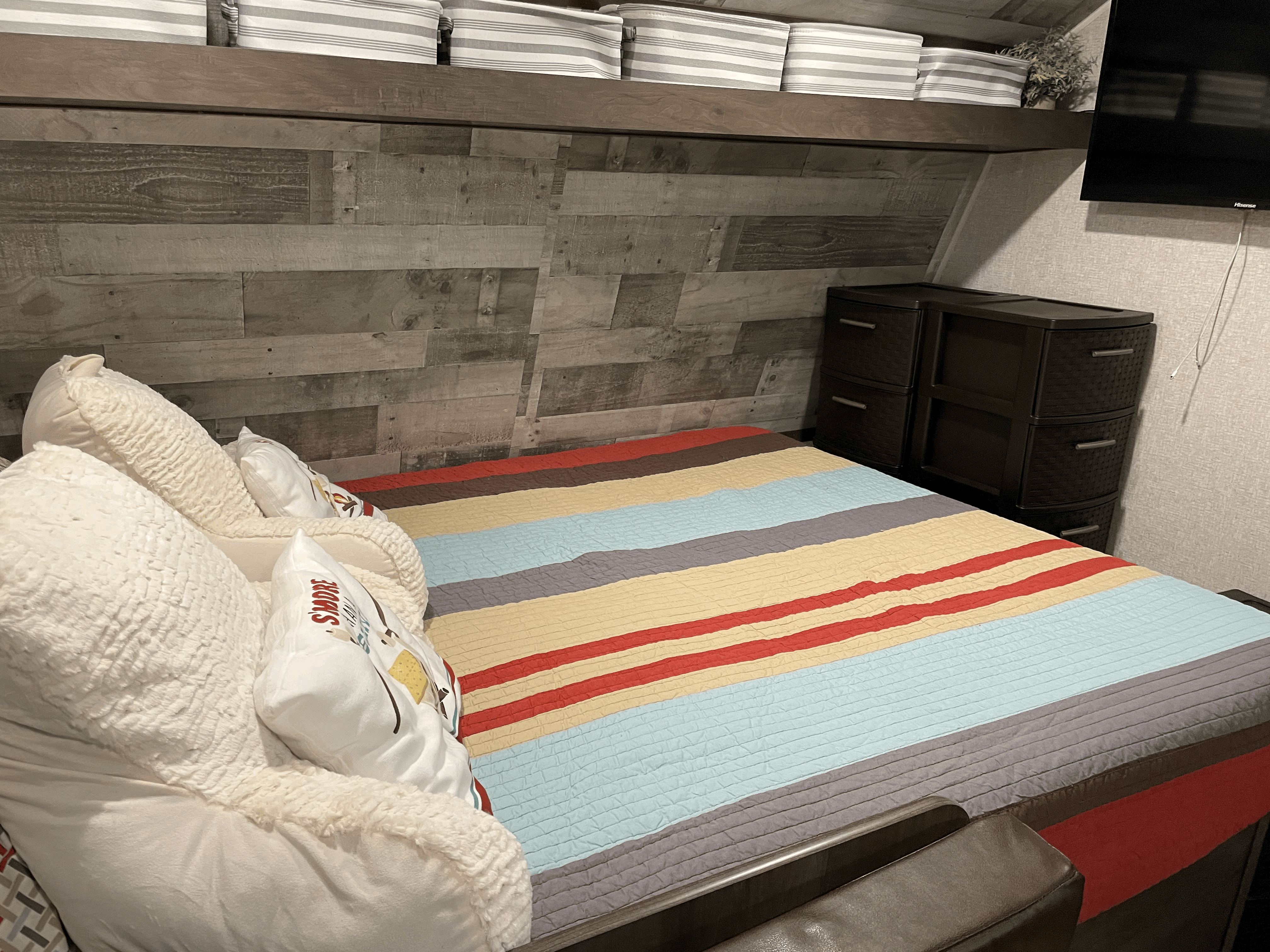 Cozy RV rental bedroom featuring a colorful striped bed, rustic wood wall, overhead storage, and dresser.. Coleman Other 2022