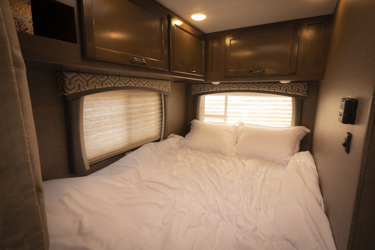 Experience comfort in this RV rental bedroom: a neat bed with white sheets, two pillows, windows with blinds, and wood cabinets.. Thor Motor Coach Chateau 2019