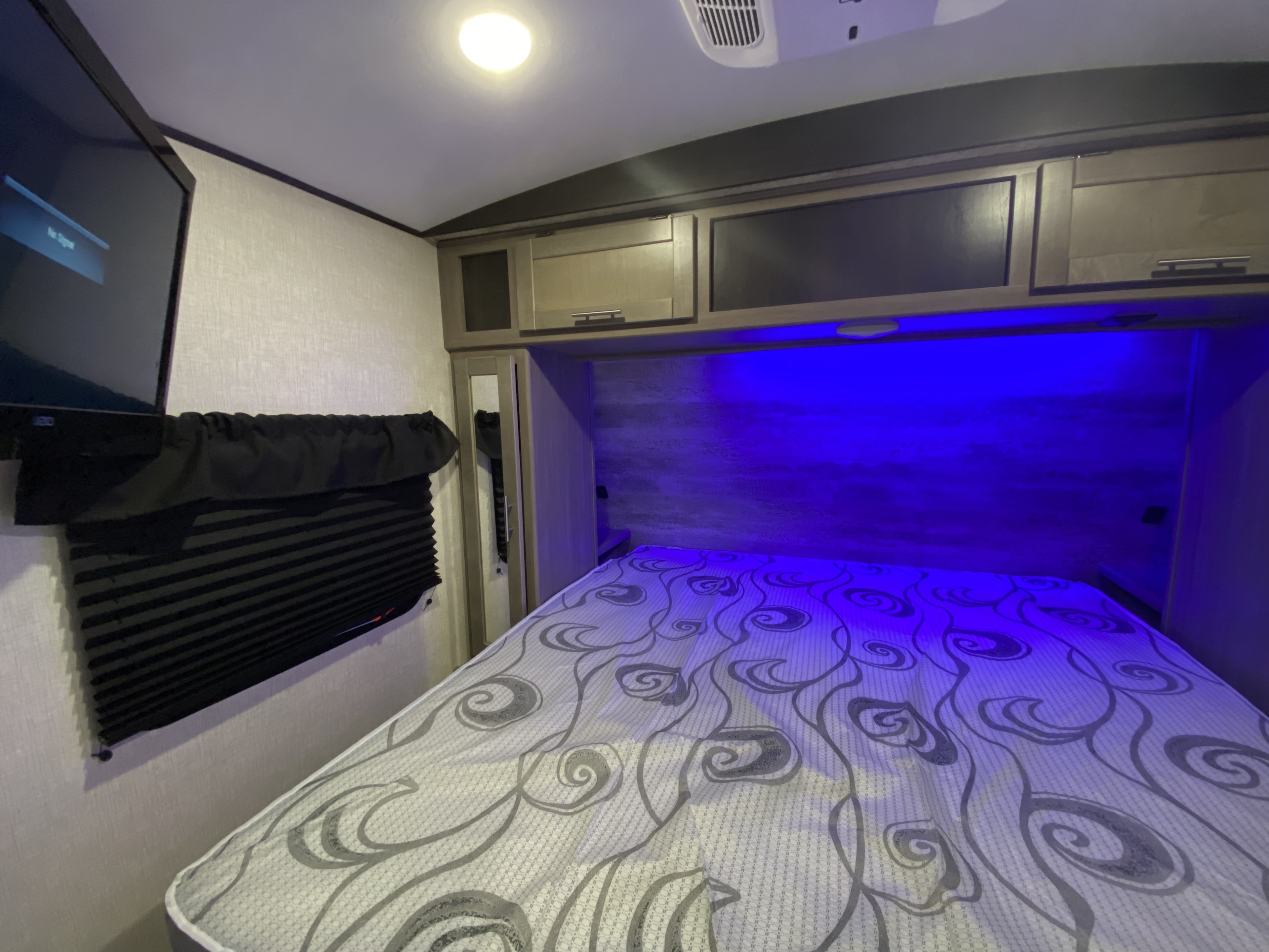Compact RV rental bedroom interior with a patterned bed, blue accent lights, overhead cabinets, TV, and window blinds.. Crossroads RV Sunset Trail Super Lite 2021