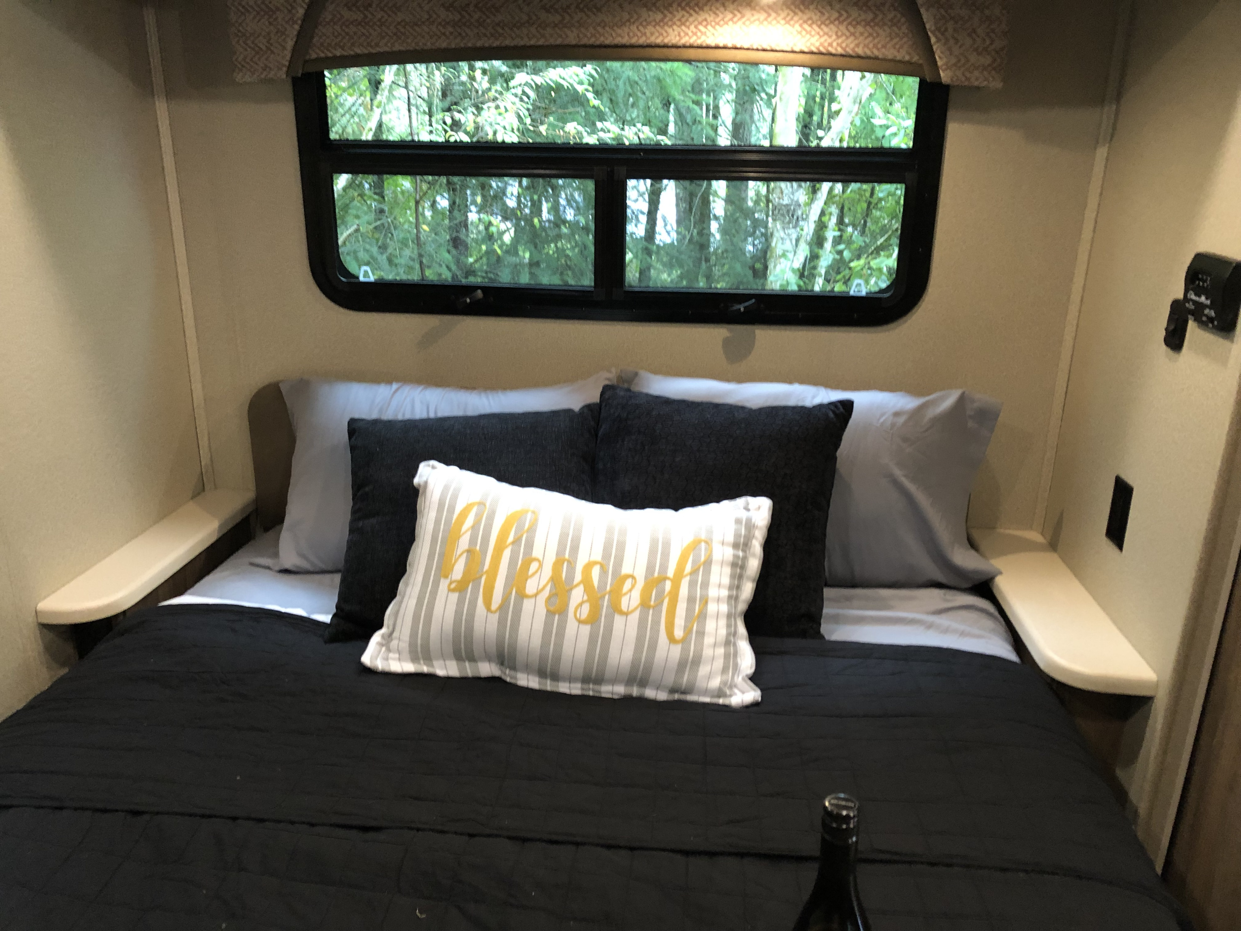 Cozy RV rental bedroom with a bed, "Blessed" pillow, and a window overlooking lush green trees, plus side shelves.. Jayco Greyhawk 2018