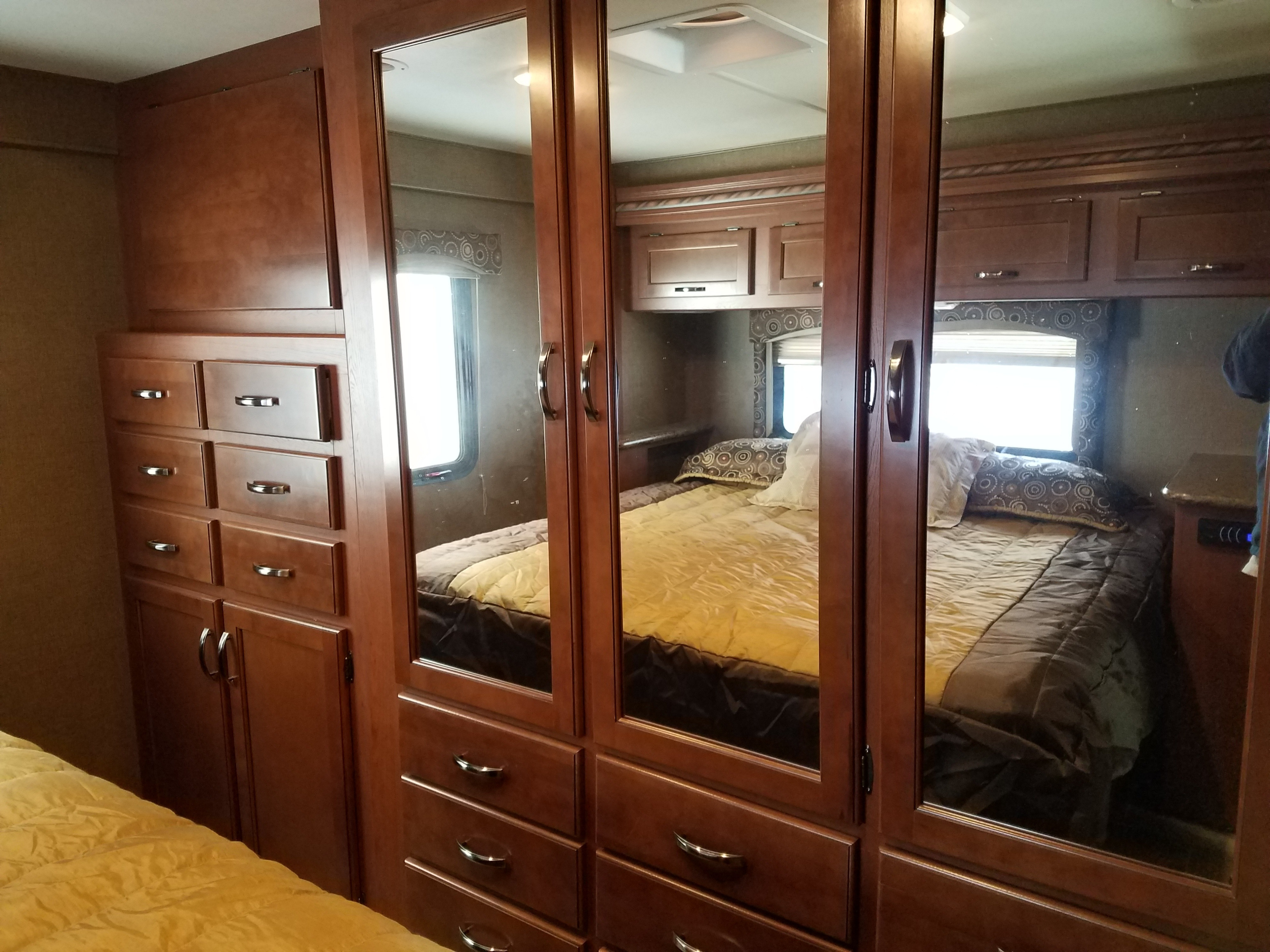 Cozy RV rental bedroom with warm wood storage, mirrored closet doors, and a comfortable bed. Mirrors reflect the room, including a window.. Thor Motor Coach Four Winds 2016