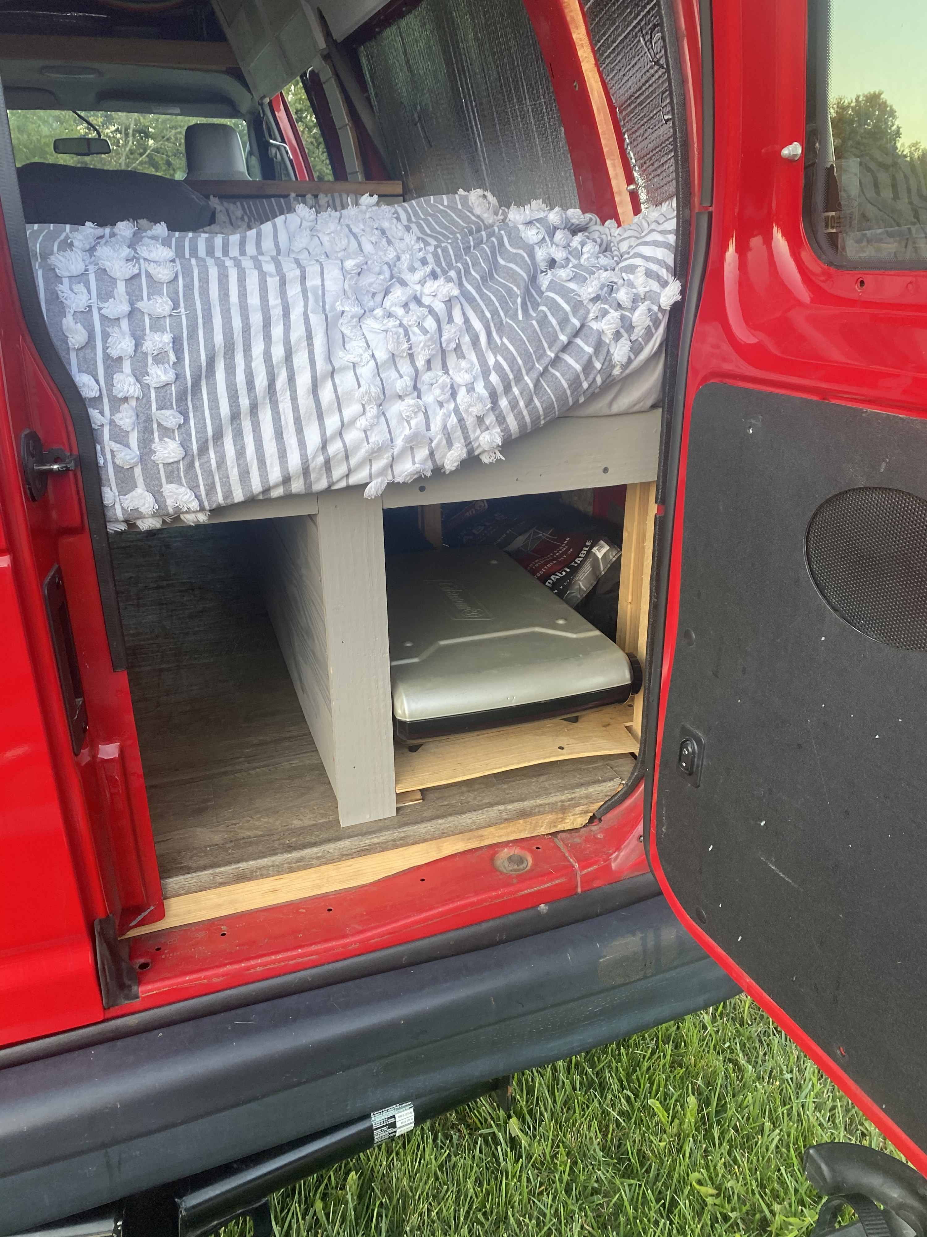 This RV rental shows a cozy van interior with a bed, under-bed storage holding a Coleman cooler, and wood flooring.. Ford Econoline E250supercab 2011