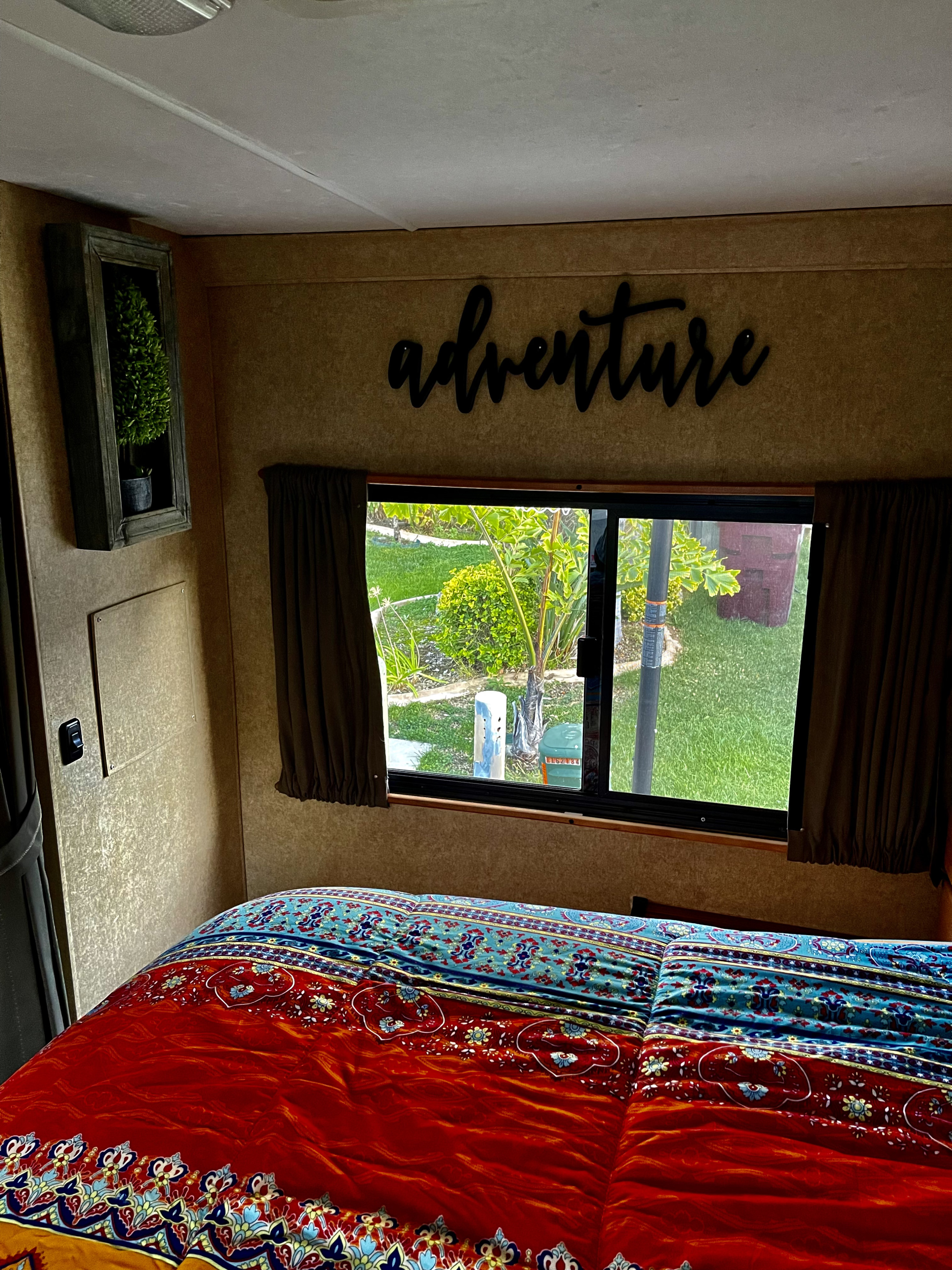 Inside an RV rental: a cozy bedroom featuring an "adventure" sign, colorful bedding, and a window view of greenery.. Other Four Winds Majestic 2017