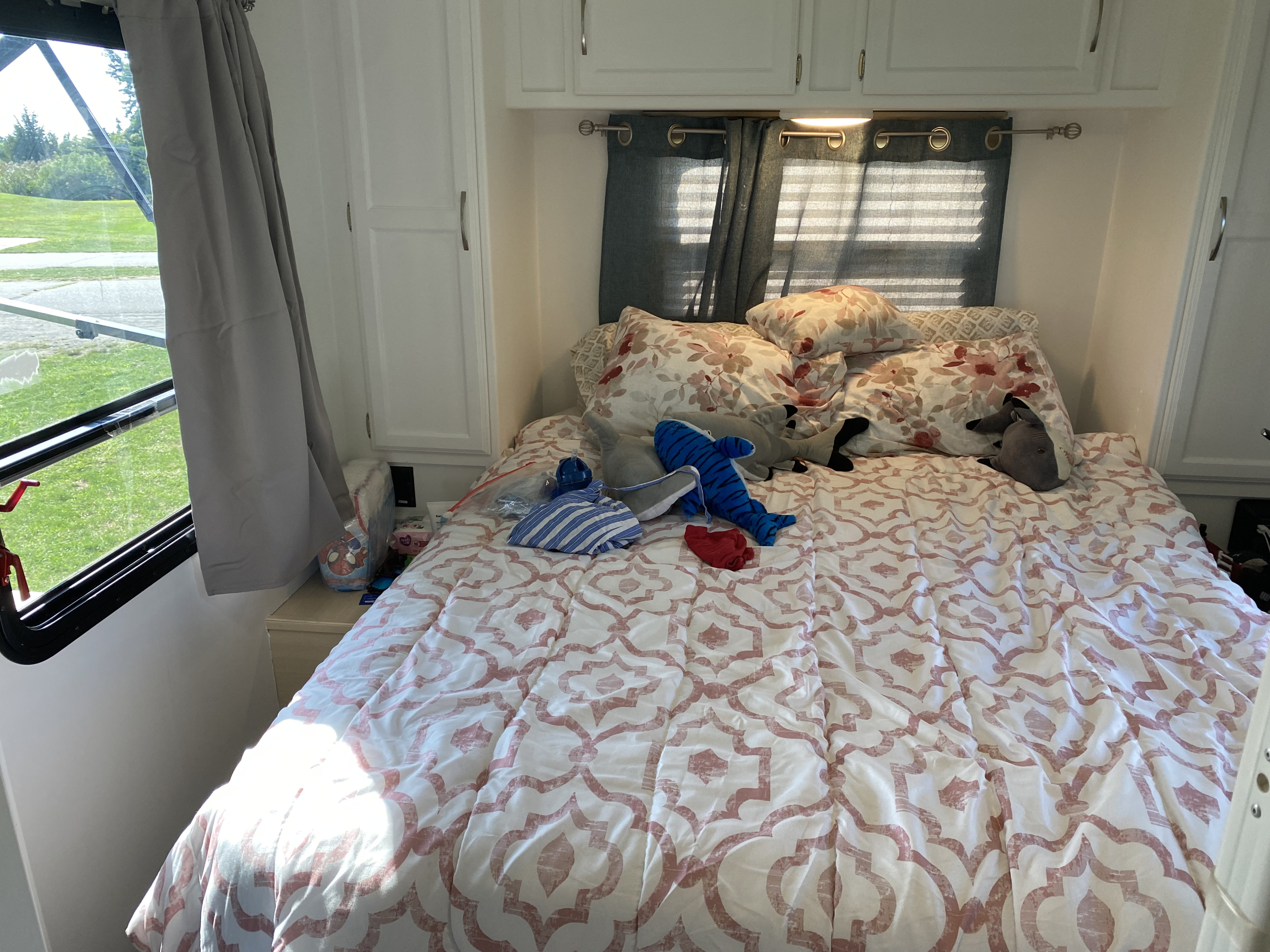 Cozy RV rental bedroom with patterned bed, floral pillows, and a bright window view of green outdoors.. Winnebago Minnie 2018
