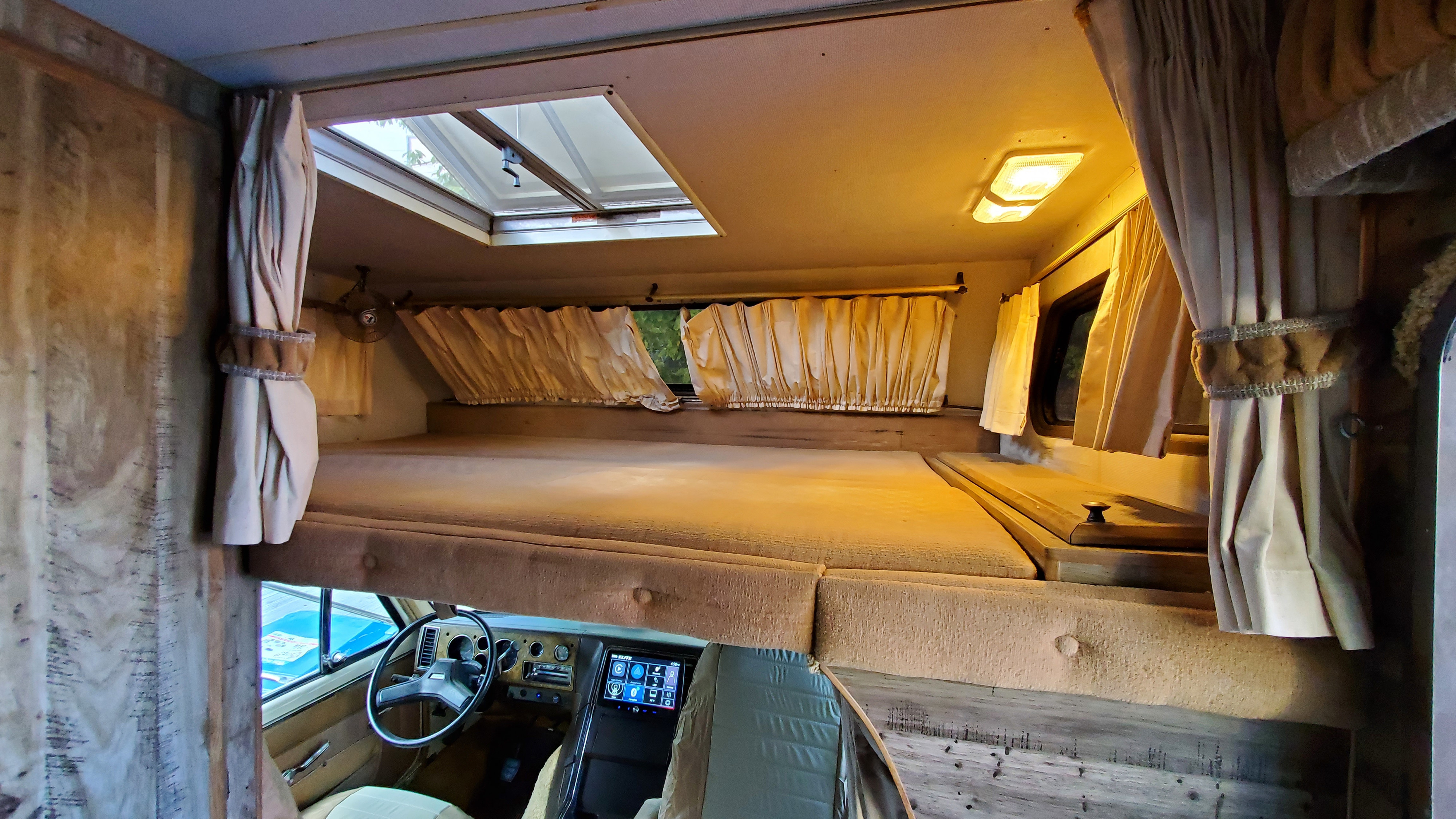 Inside an RV rental: a spacious cab-over bunk with skylight, windows, curtains, and the driver's area below.. Lazy Daze 22 1987