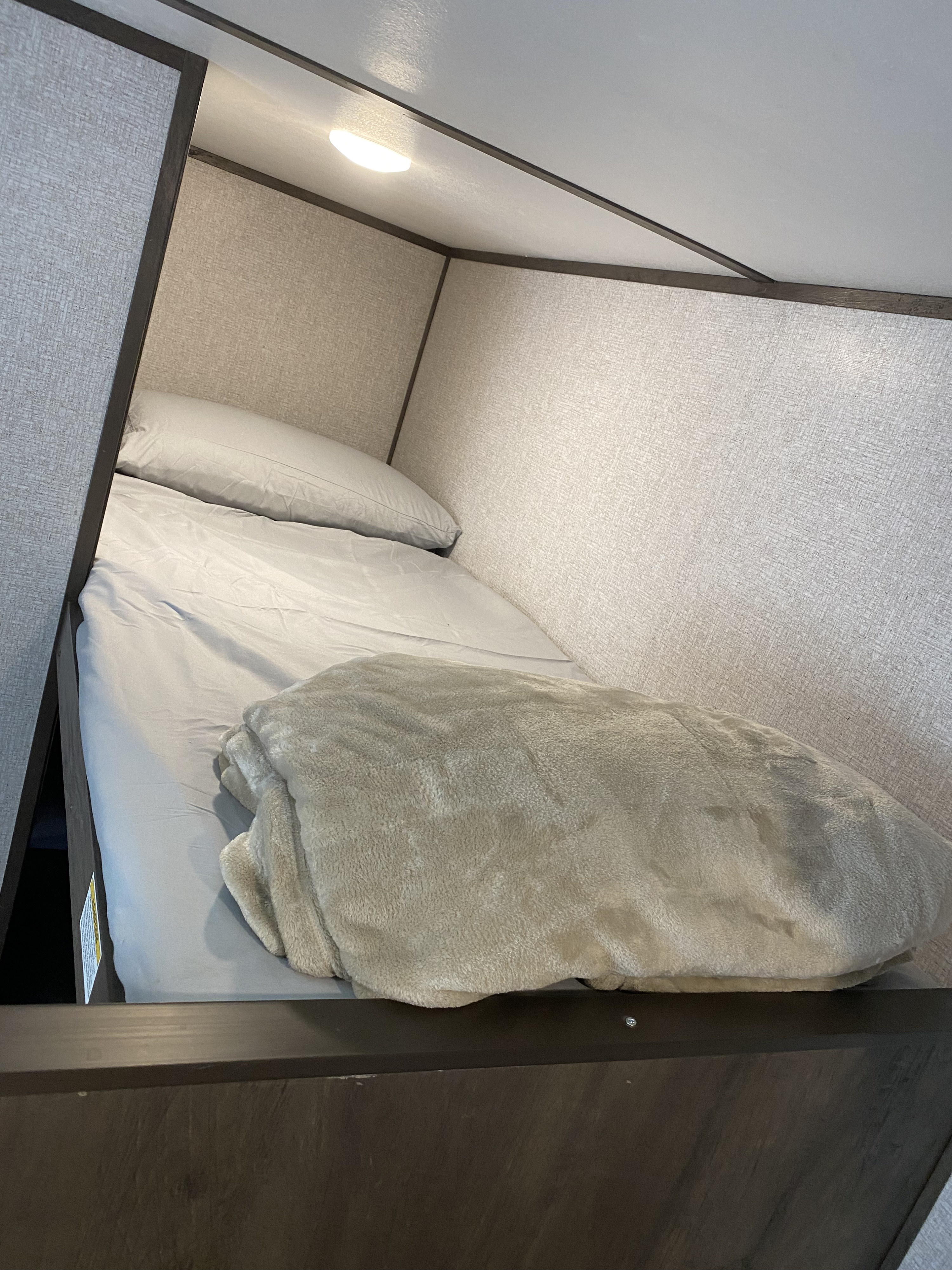 Inside an RV rental: a neat bunk bed with light bedding, a pillow, and a cozy blanket, lit by an overhead light.. Coleman Lantern 17 B 2022