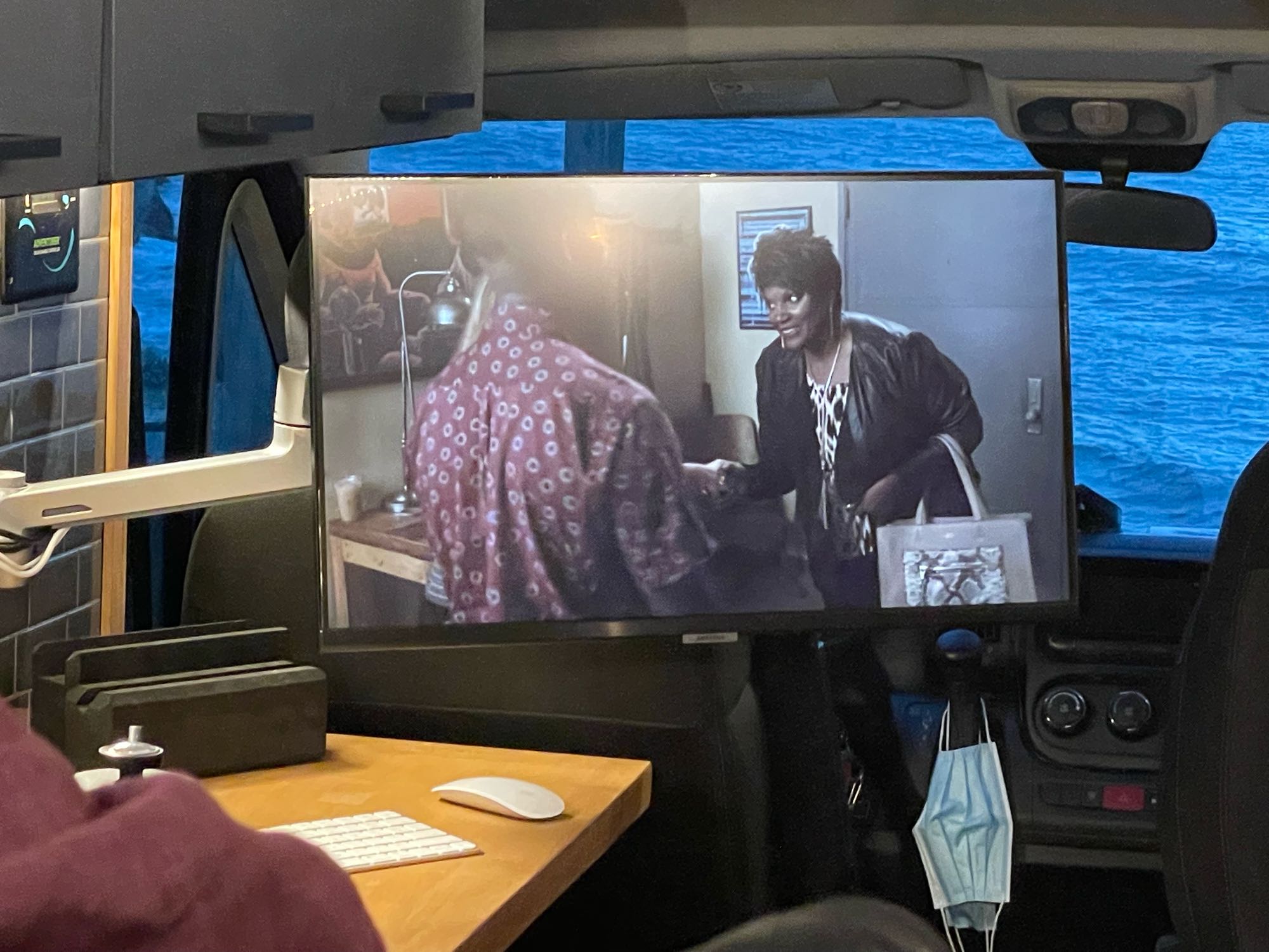 Inside an RV rental: a mobile workspace with a large screen for entertainment, overlooking calming blue water.. Ram Promaster 2021