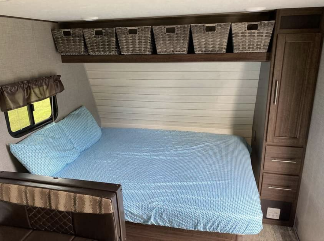 Inside an RV rental bedroom, featuring a blue-sheeted bed, overhead storage baskets, window, and wood wardrobe.. Crossroads Zinger 2018