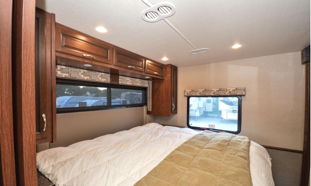 Interior view of an RV rental bedroom showing a bed with white and tan covers, wooden cabinets, and windows.. Fleetwood Other 2017