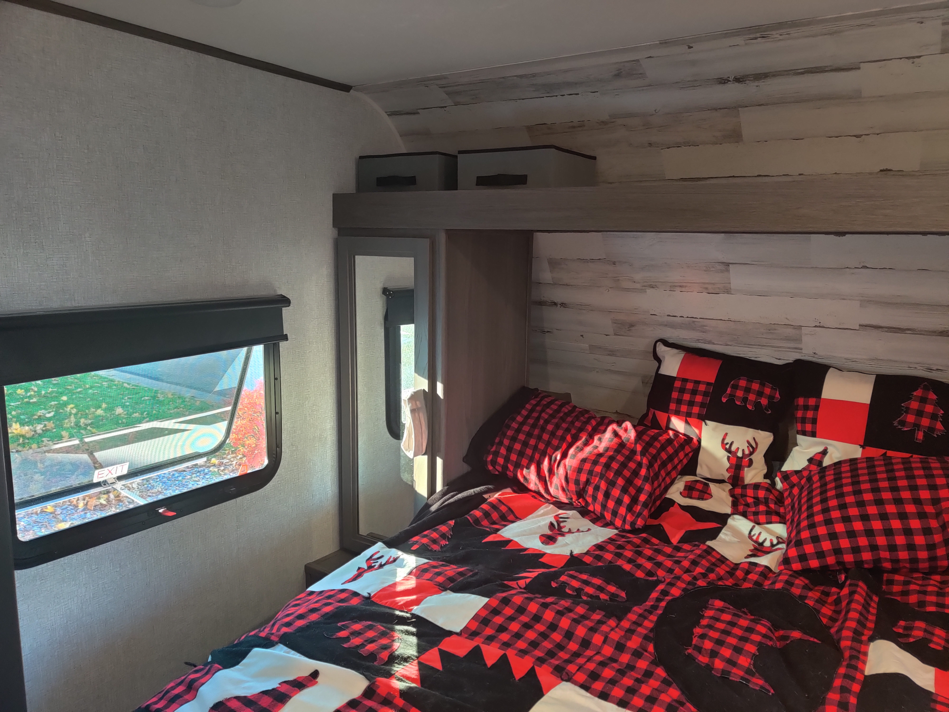 Cozy RV rental bedroom with red & black buffalo plaid bedding. Features deer, bear, tree designs, a window with outdoor view, and storage.. Dutchmen Kodiak 2021