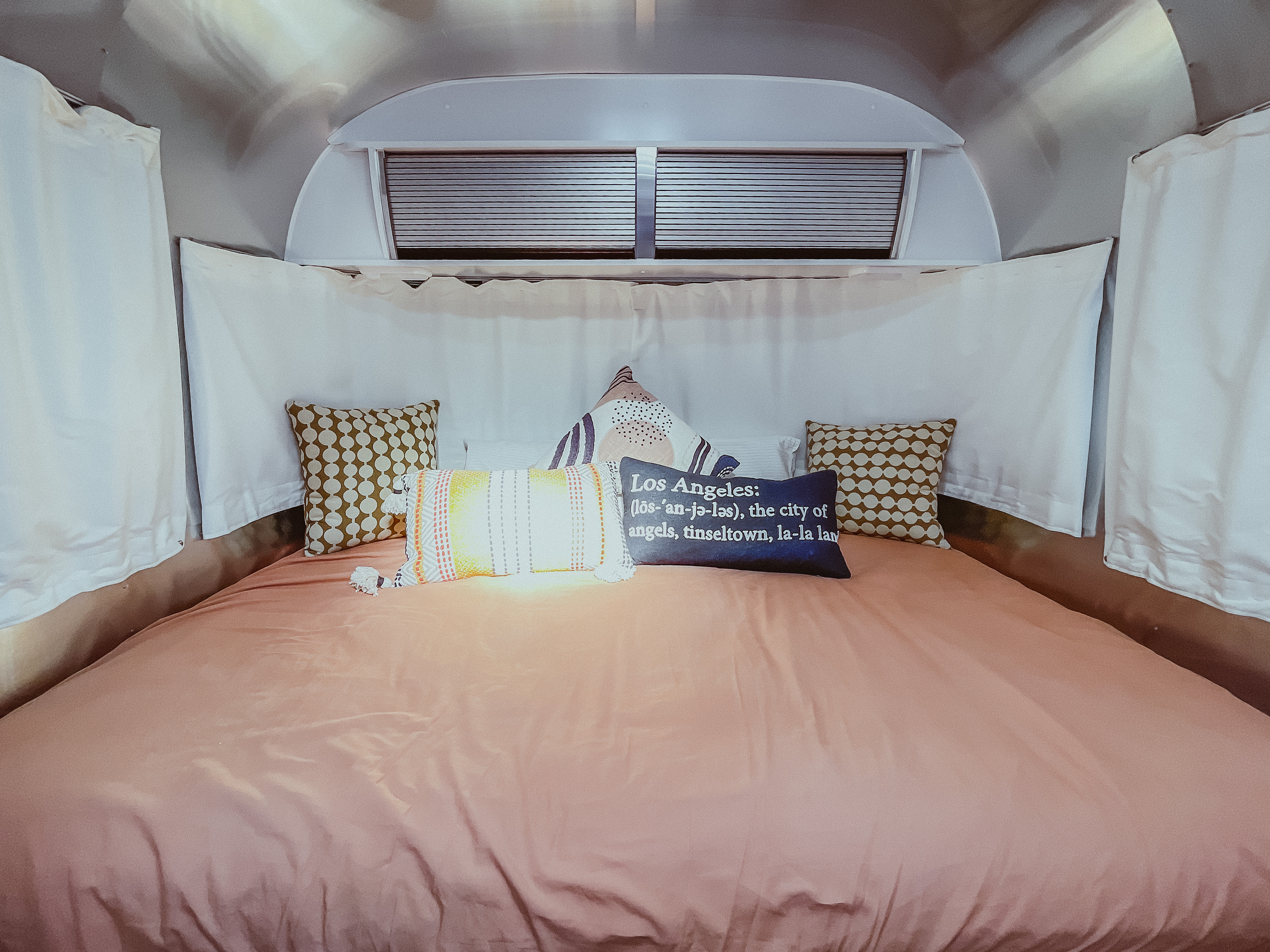 Cozy RV rental bedroom with peach bedding and a Los Angeles pillow, offering a comfortable space for travel.. Airstream Sport 2019