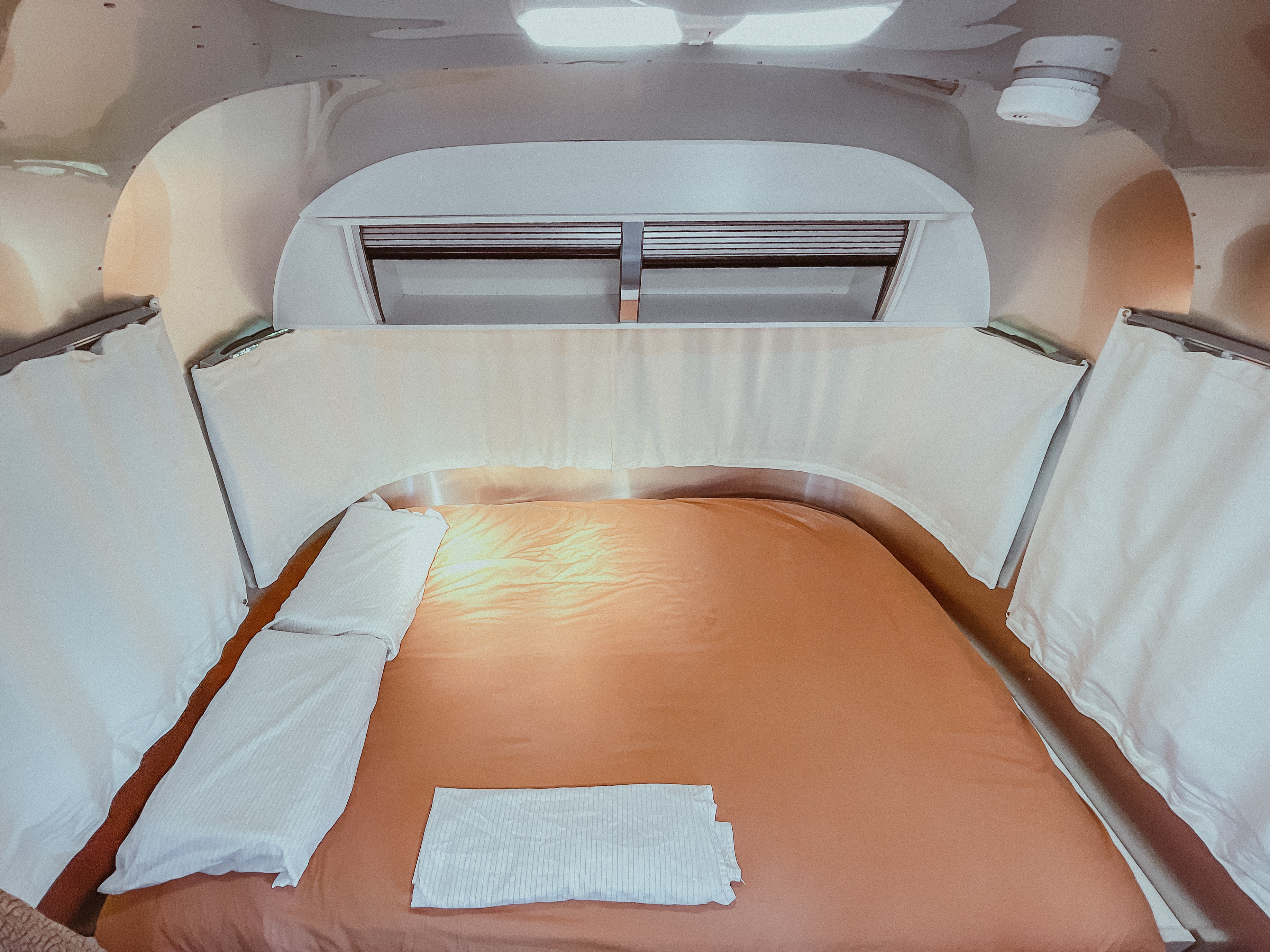 Cozy RV rental bedroom featuring a bed with warm orange sheets, white linens, and overhead storage.. Airstream Sport 2019
