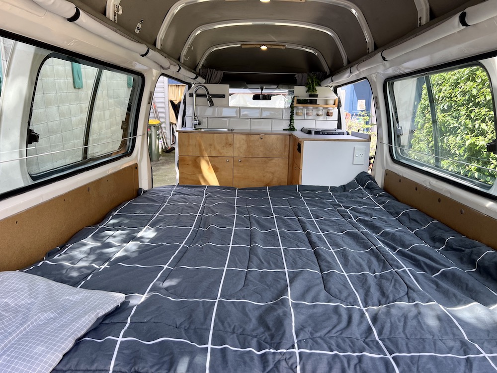 Inside a modern RV rental: comfy bed with grid comforter, full kitchen setup & natural light.. Nissan Caravan 2005