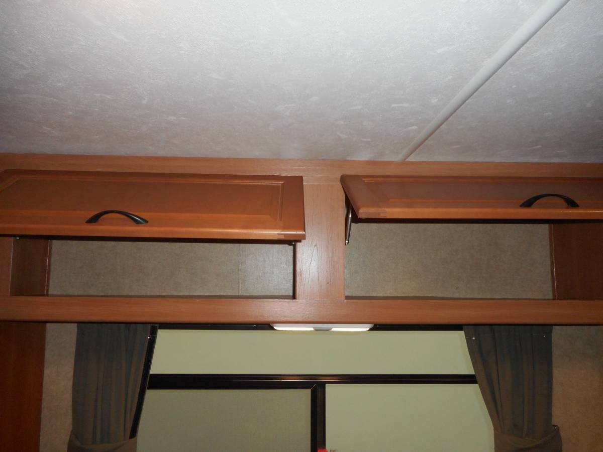 An RV rental interior features overhead storage cabinets, one open, above a window with curtains and lighting.. Thor Majestic 2017