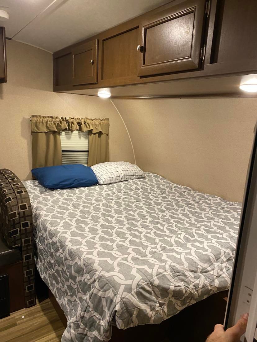 Comfortable RV rental bedroom: features a full bed with patterned bedding, overhead cabinets, and a window.. Shasta Oasis 18BH 2018