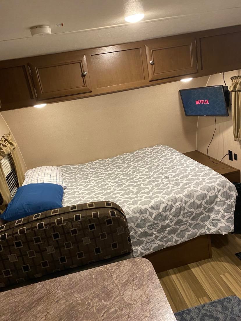 This RV rental offers a cozy bedroom with a patterned bed, mounted TV, overhead cabinets, and a dinette.. Shasta Oasis 18BH 2018