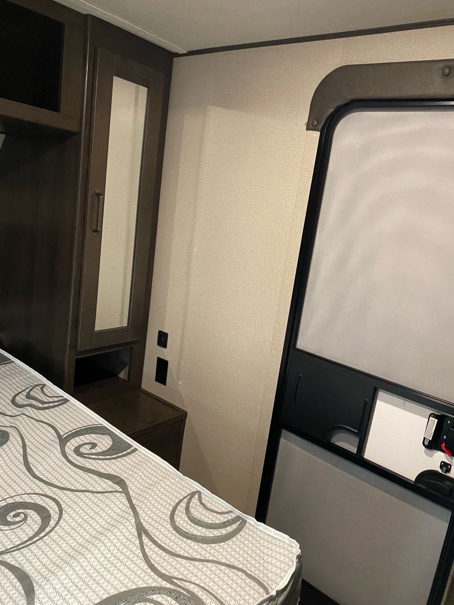 Inside an RV rental: a compact sleeping area with patterned mattress, dark wood cabinets including a mirrored door, and the entrance.. Grand Design Transcend 2022