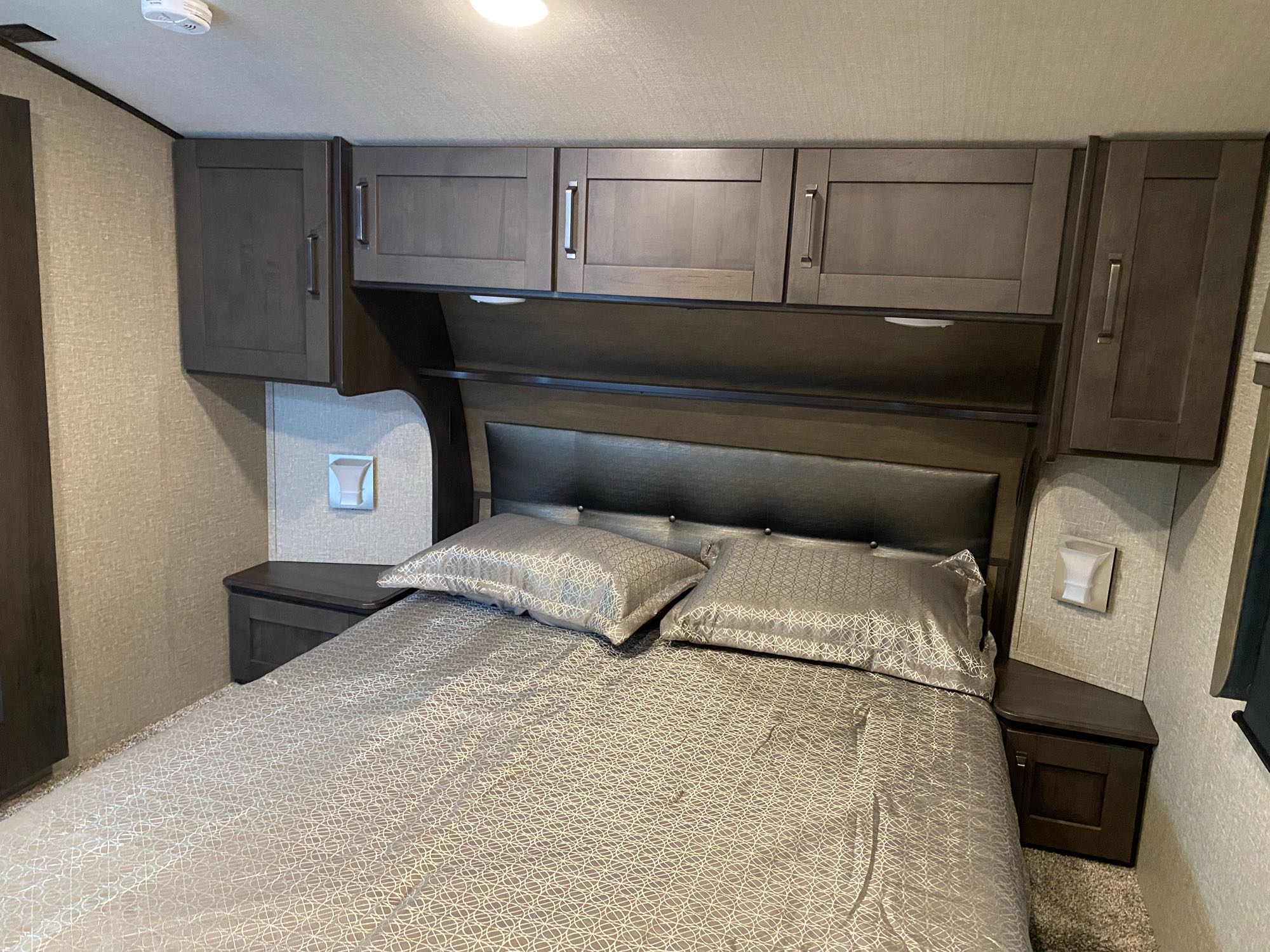 Inside an RV rental: a neat bedroom with a made bed, patterned bedding, dark wood cabinets, and built-in bedside storage.. Grand Design Reflection 2022