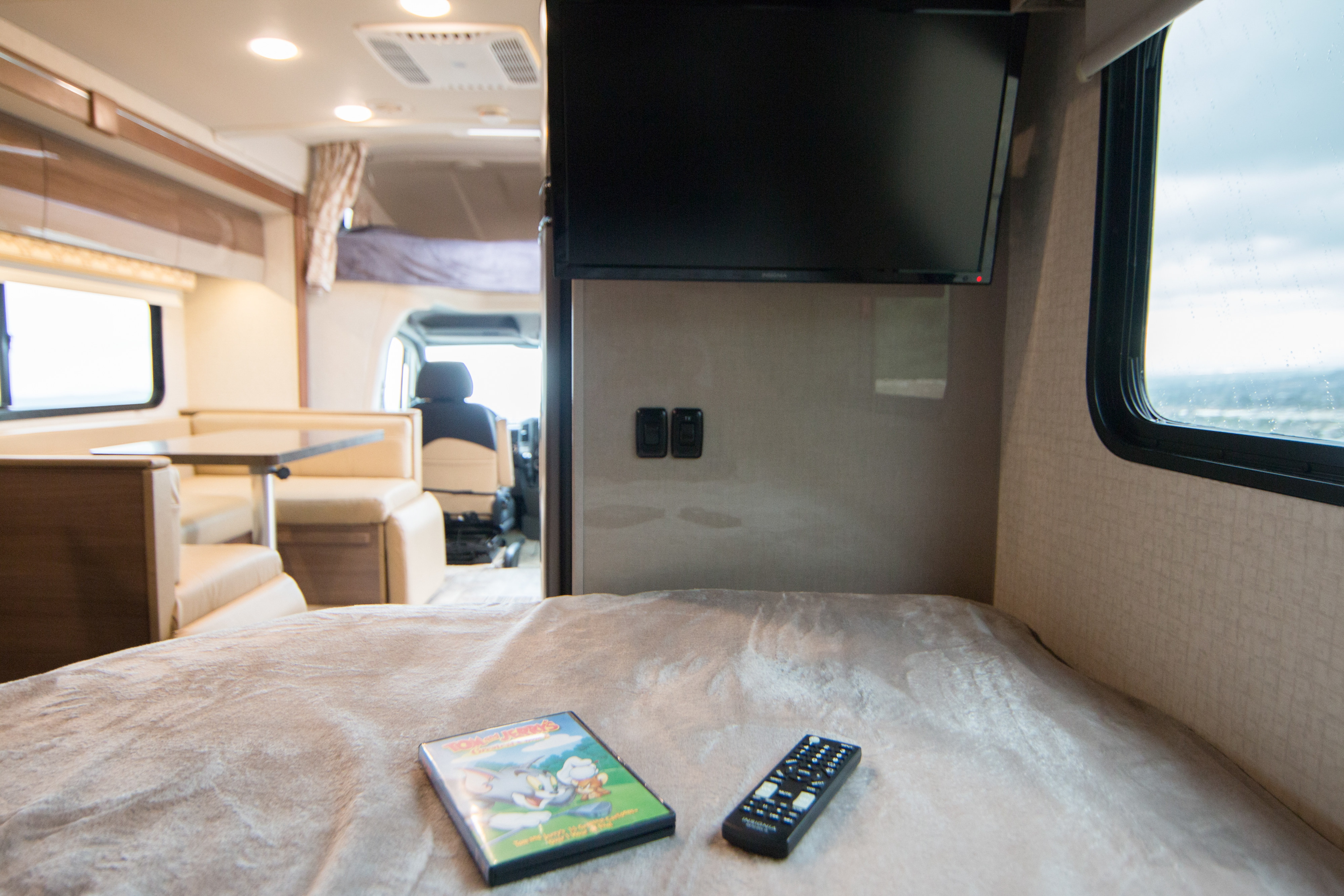 RV rental interior: a comfy bed with a Tom & Jerry DVD and remote, a mounted TV, and a dining area in view.. Itasca Navion 2017