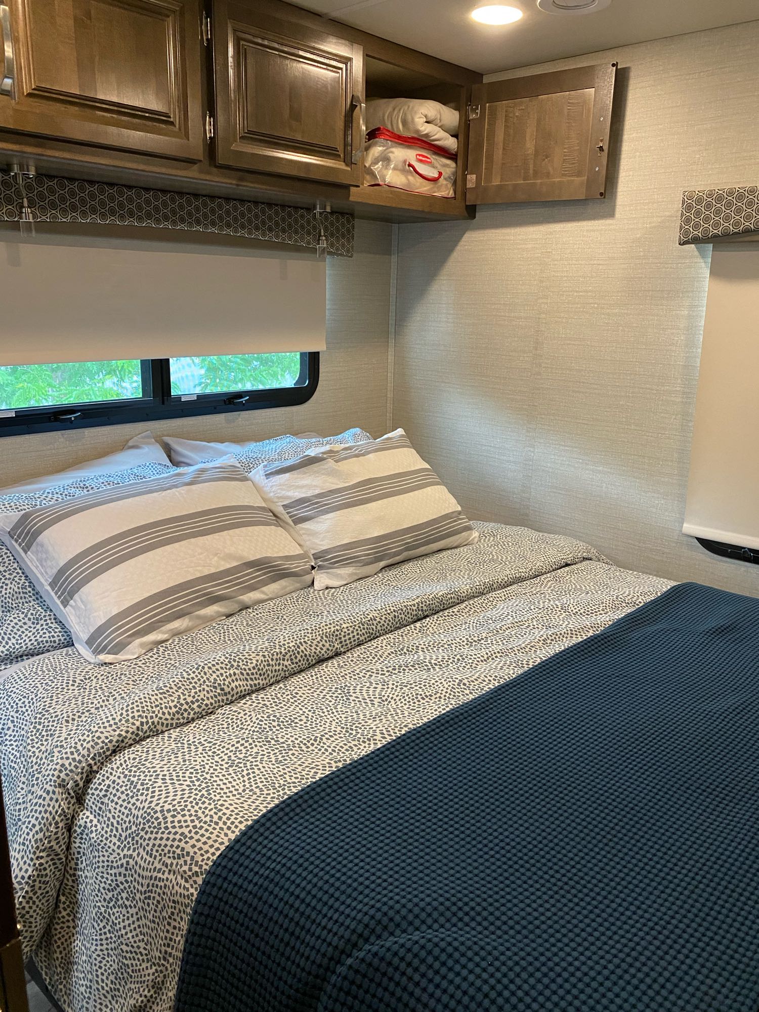 Cozy RV rental bedroom features a comfy bed with striped pillows, patterned bedding, open overhead cabinet, and windows.. Jayco Greyhawk 2019