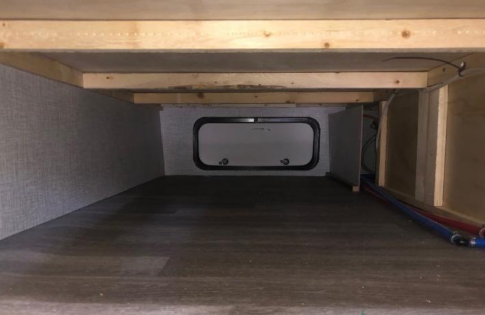RV rental under-bed storage compartment, featuring a central access hatch, wood floor, and visible plumbing.. Braxton Creek Bushwhacker Plus 2021