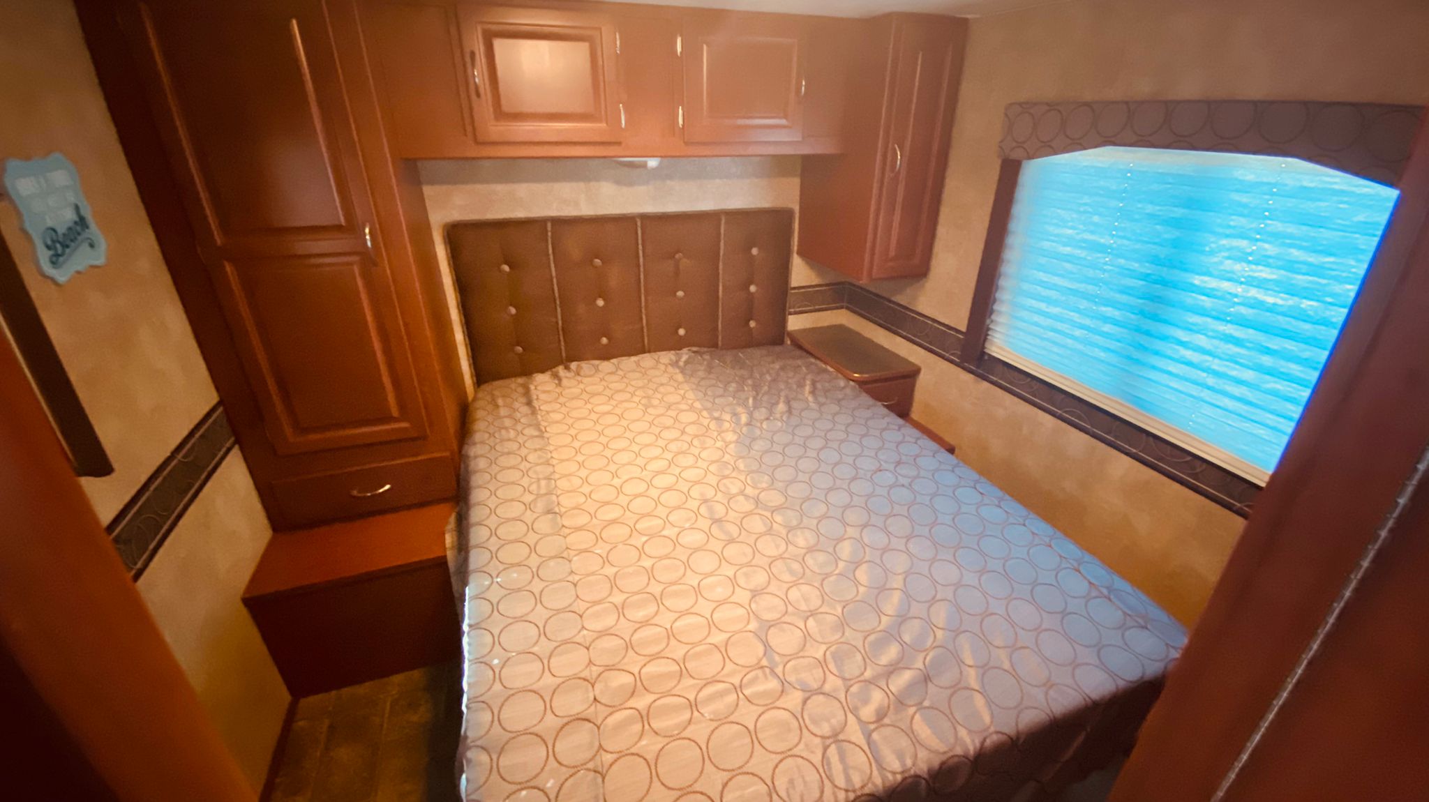 Inside an RV rental, a compact bedroom features a comfortable bed, wooden storage cabinets, and a window with blue blinds.. Winnebago 31k 2015