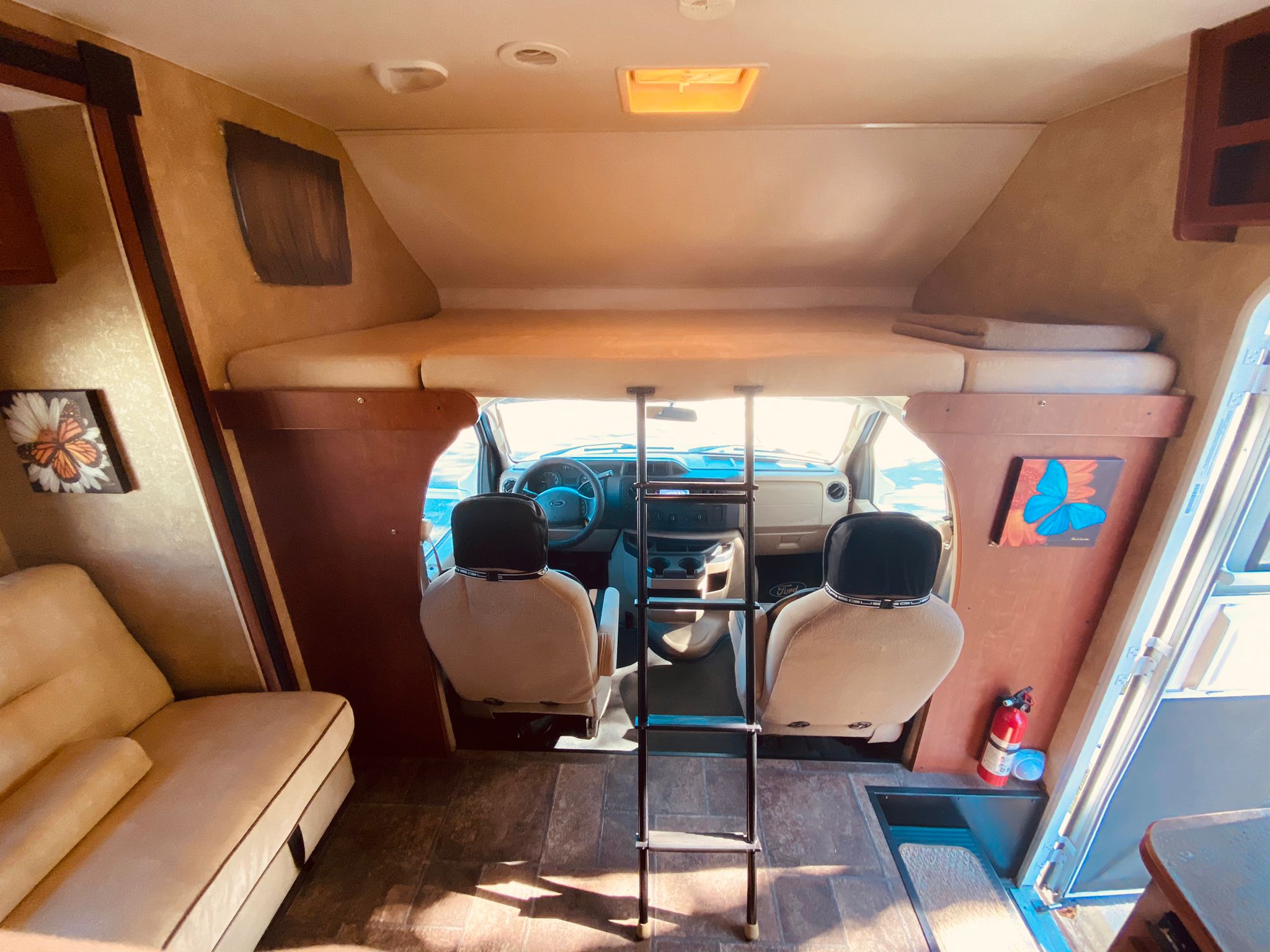 Bright RV rental interior: overhead bed with ladder, cockpit, and lounge seating. Fire extinguisher on the right.. Winnebago 31k 2015